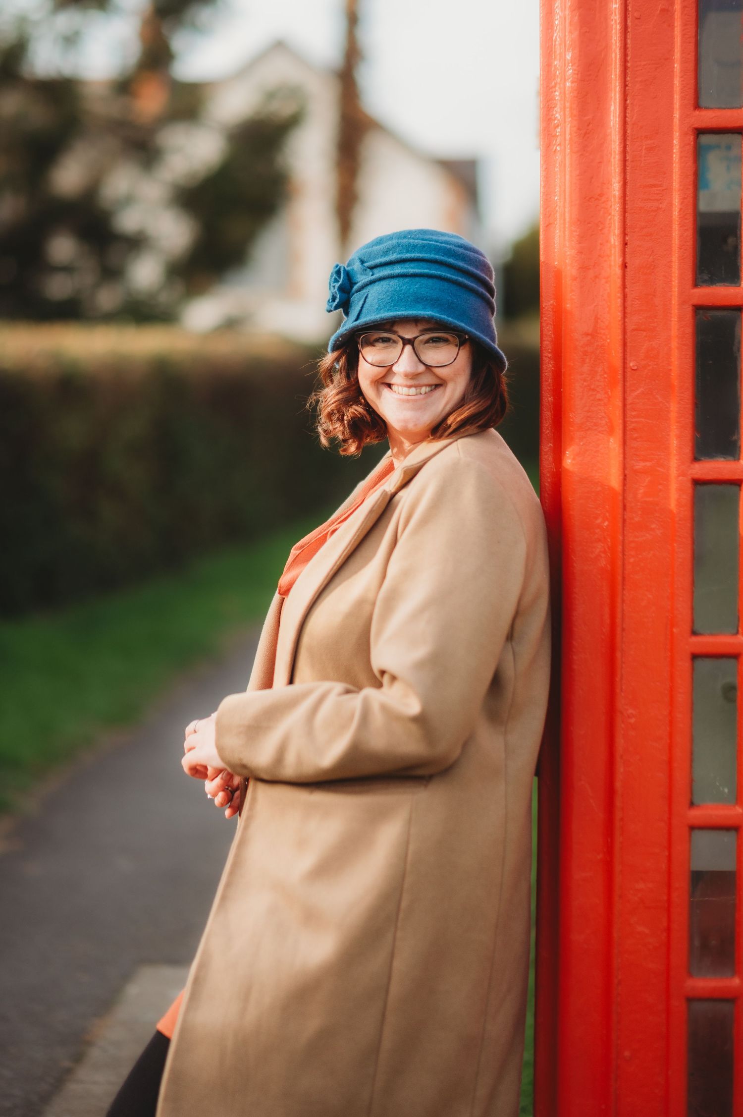 Creative marketing consultant standing beside a red telephone box, smiling and wearing glasses, a blue hat and a coat.