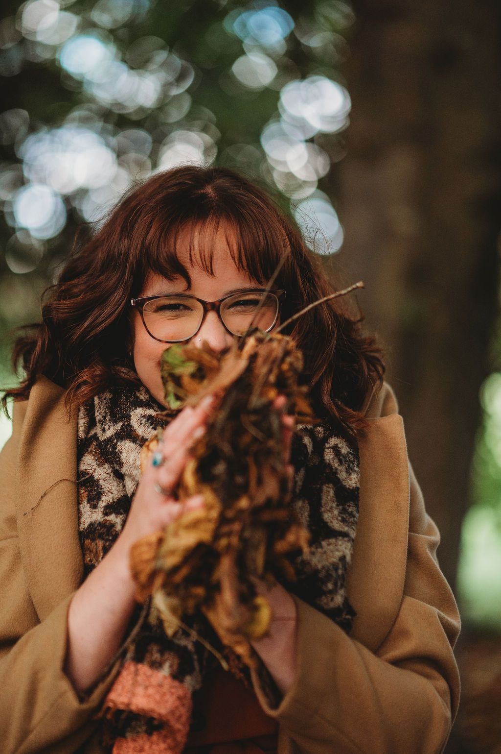 Creative marketing consultant smiling outdoors while holding a handful of autumn leaves, wearing glasses and a patterned scarf.