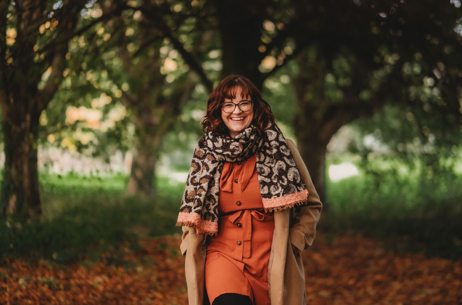 Creative marketing consultant standing outdoors among autumn leaves, smiling and wearing glasses, a scarf and a coat.