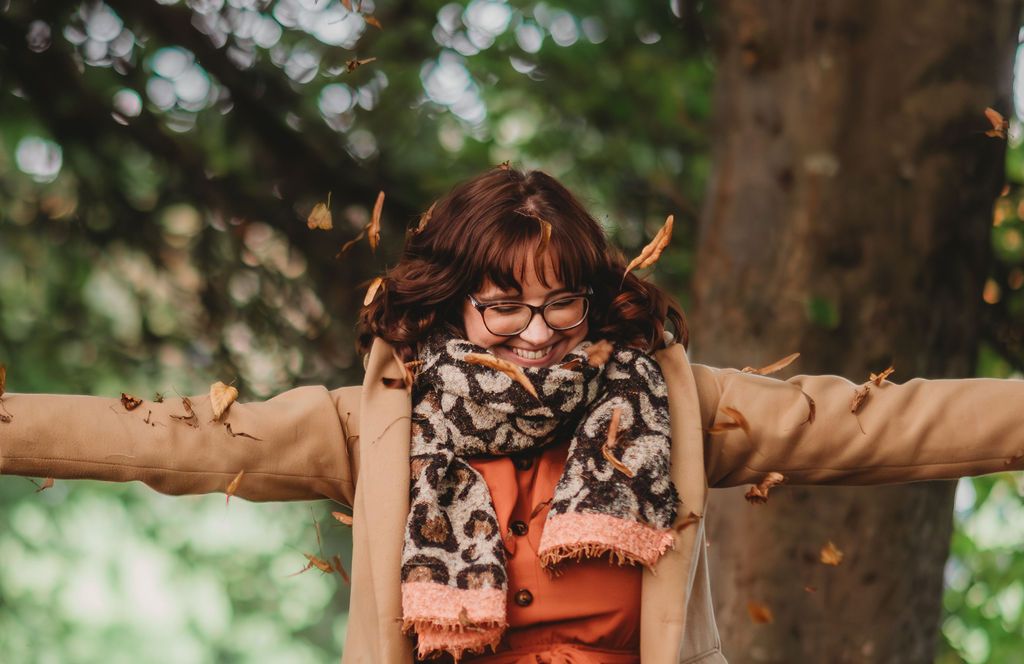 Founder of Alba & Bright enjoying an outdoor moment with arms outstretched, wearing a scarf and glasses beneath trees.