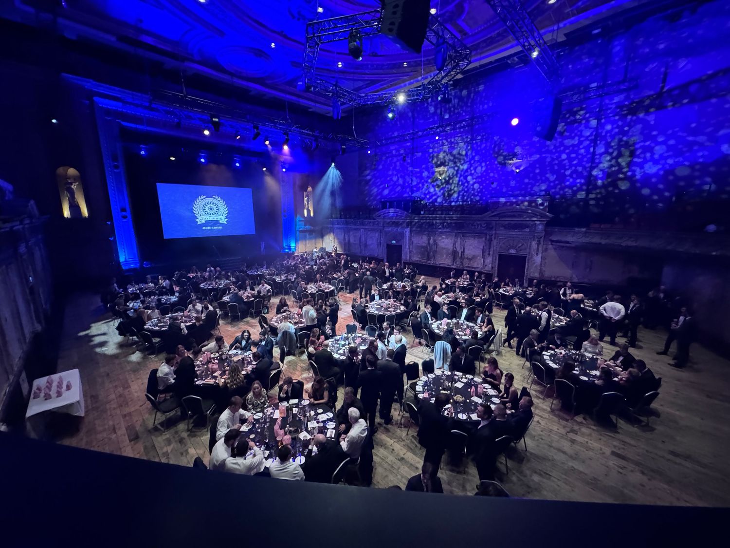 Large banquet hall illuminated in blue lighting with multiple round tables and guests seated for a formal event.