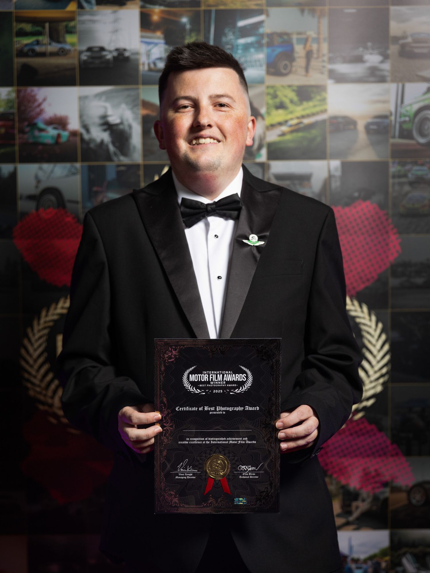 A person in a black tuxedo and bow tie holds up an award certificate in a formal setting.