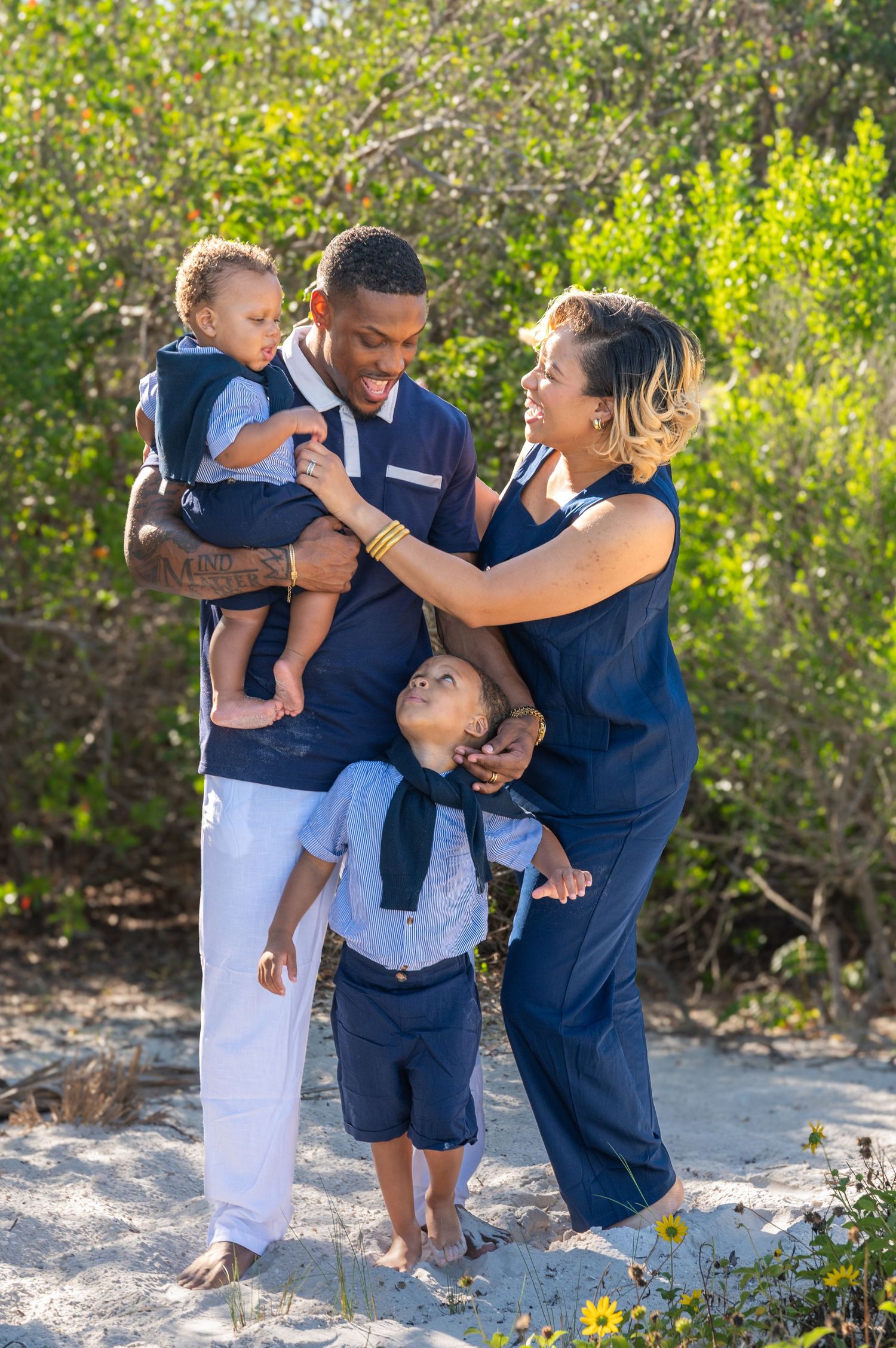 Happy family of four in navy outfits posing outdoors on sandy beach with lush green foliage background.
