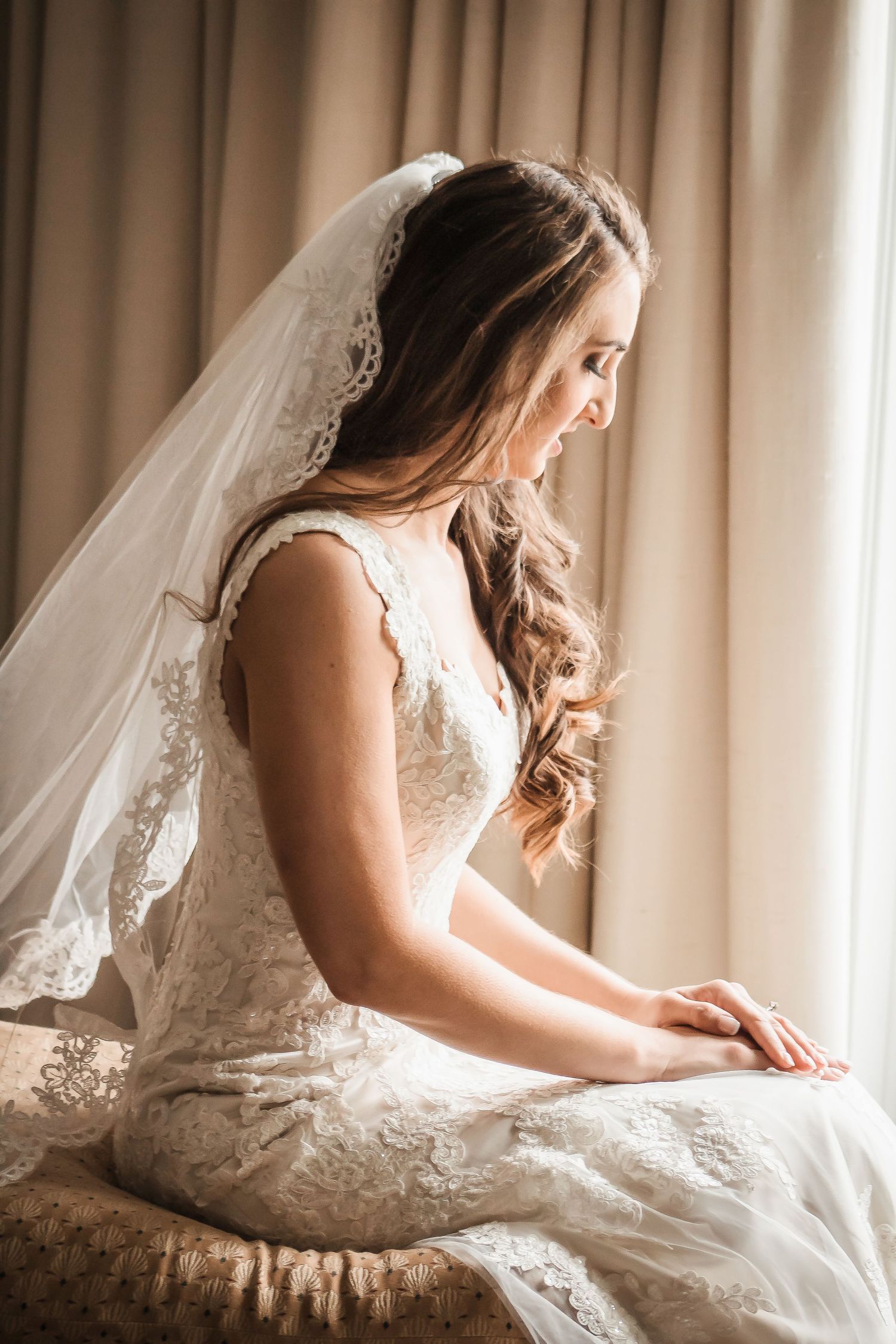 Bride in lace wedding dress and veil sits gracefully by a sunlit window, looking down with a serene expression.