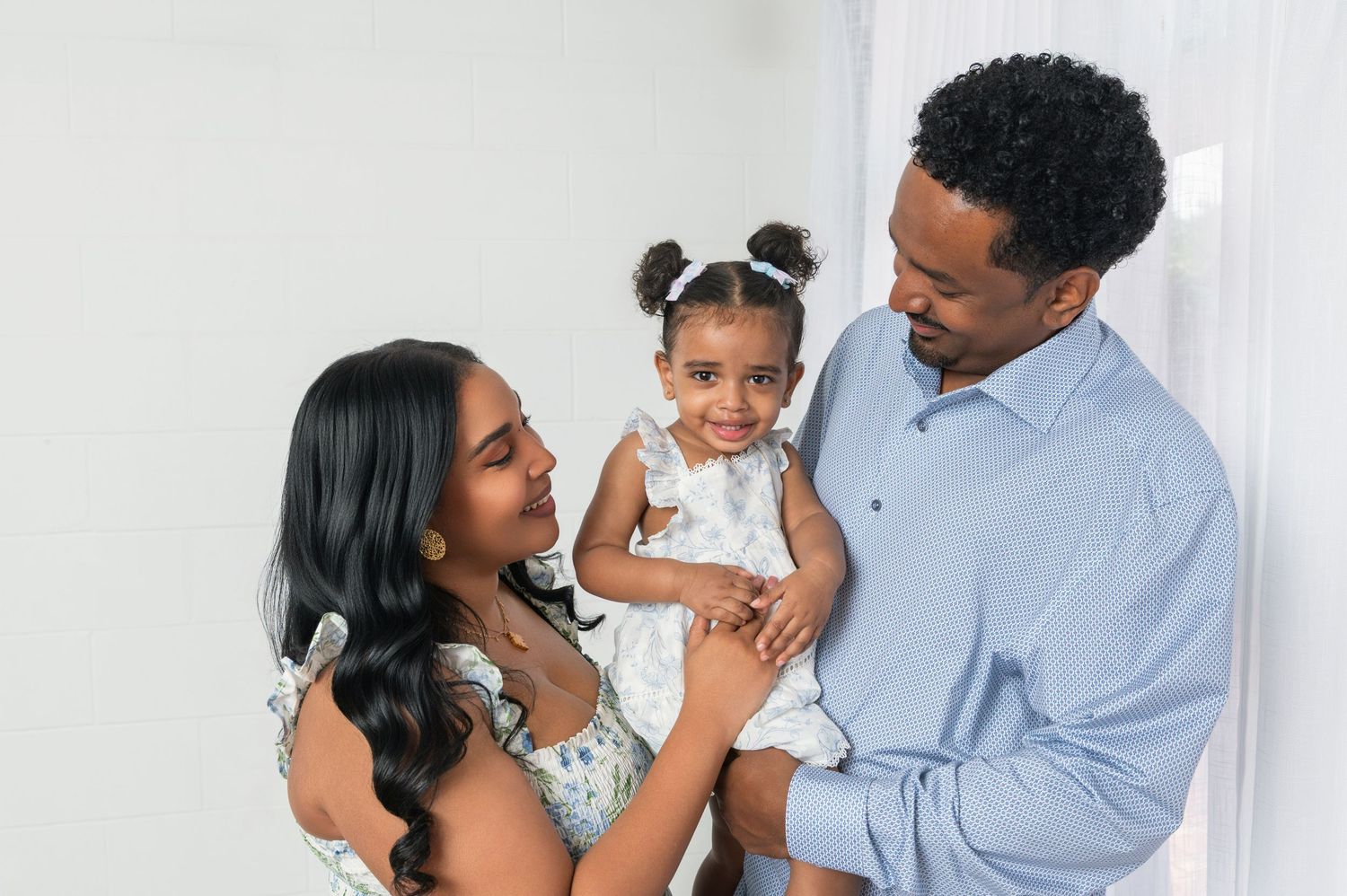 A happy Black family with parents smiling at their toddler daughter in pigtails during a professional photo session.