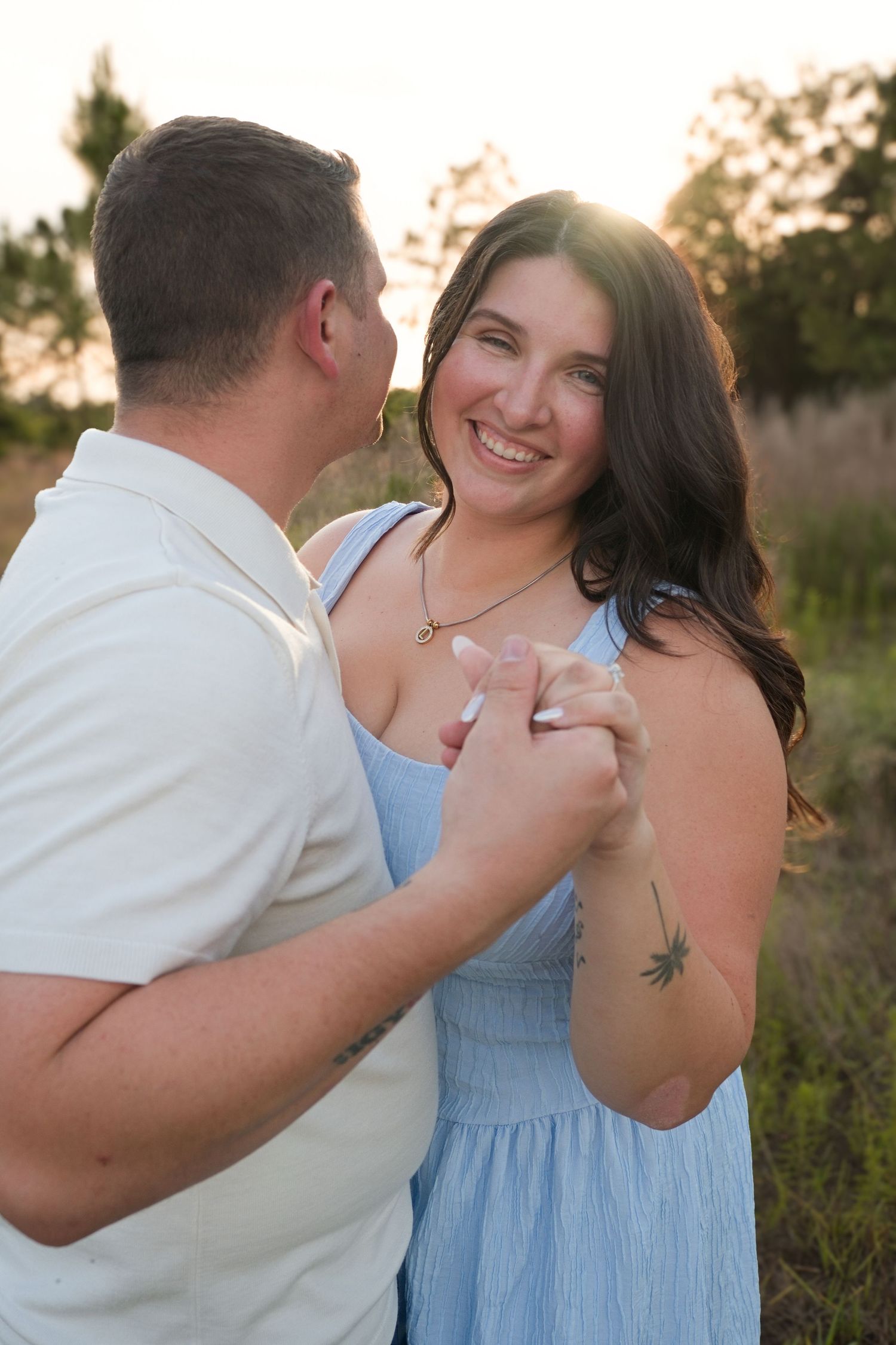 A smiling woman in a blue dress holds hands with a man outdoors at golden hour in a field.
