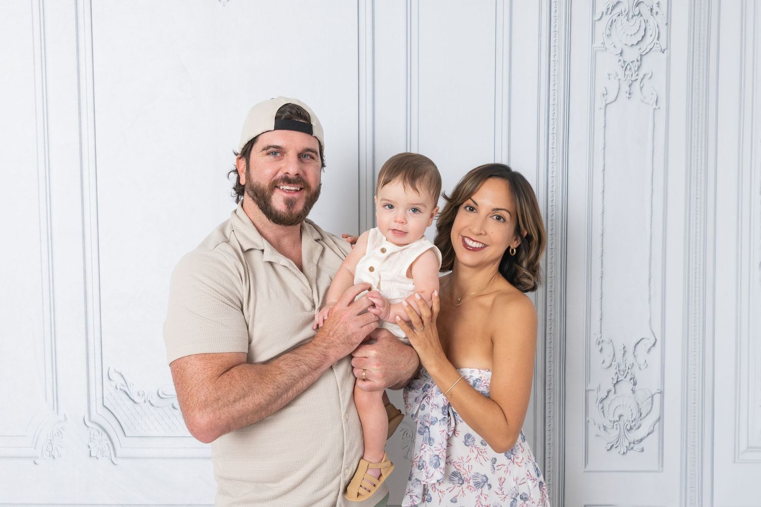 A smiling couple poses with their baby in a bright white studio setting, the man wearing a backwards cap.