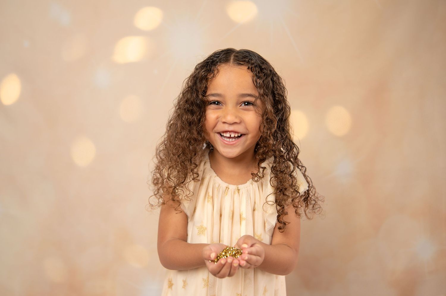 Smiling young girl with curly hair in a cream dress holding glitter against a bokeh light background.