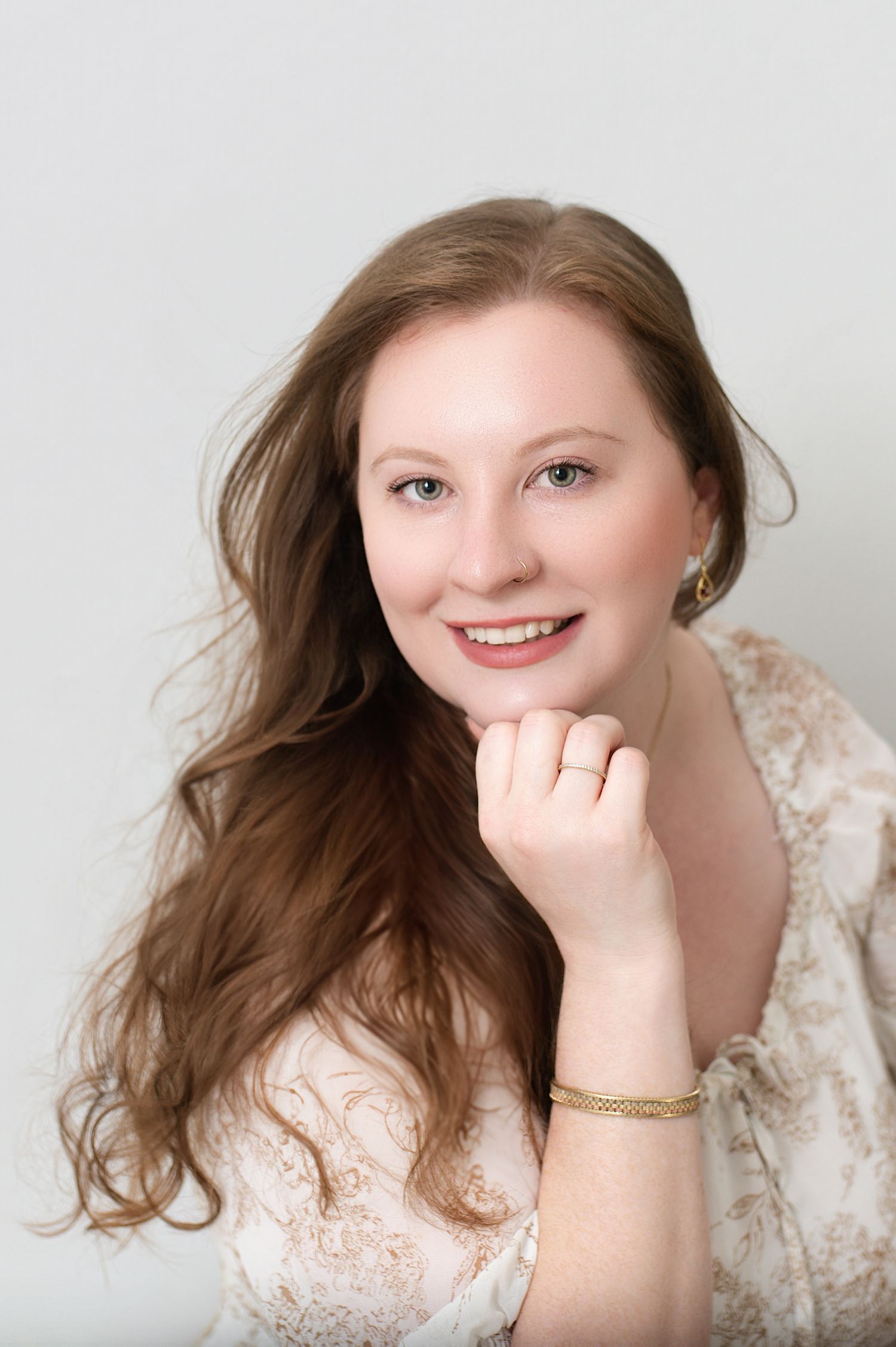 Portrait of someone with long brown hair wearing a lace dress and jewelry against a light background.