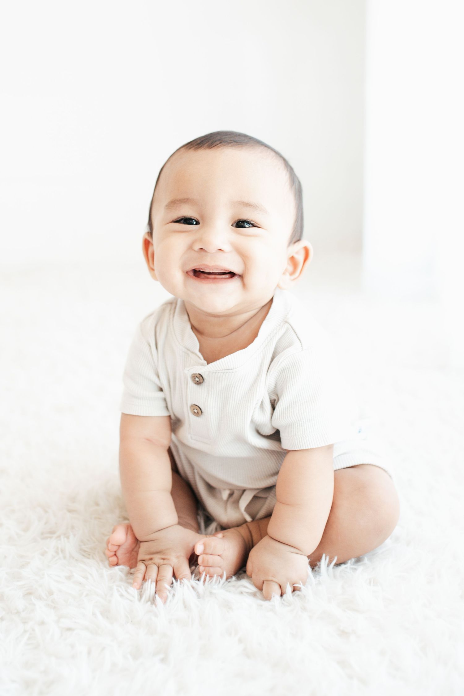 Smiling baby in white onesie crawls on fluffy white carpet during bright studio photoshoot.