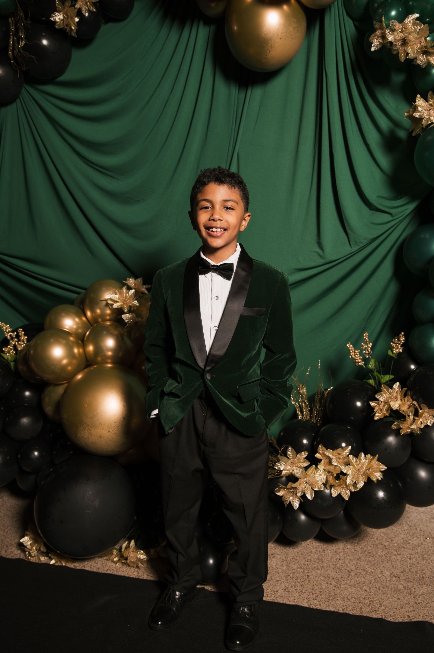 A smiling boy in a green velvet tuxedo with bow tie poses in front of a green backdrop with gold and black balloons.