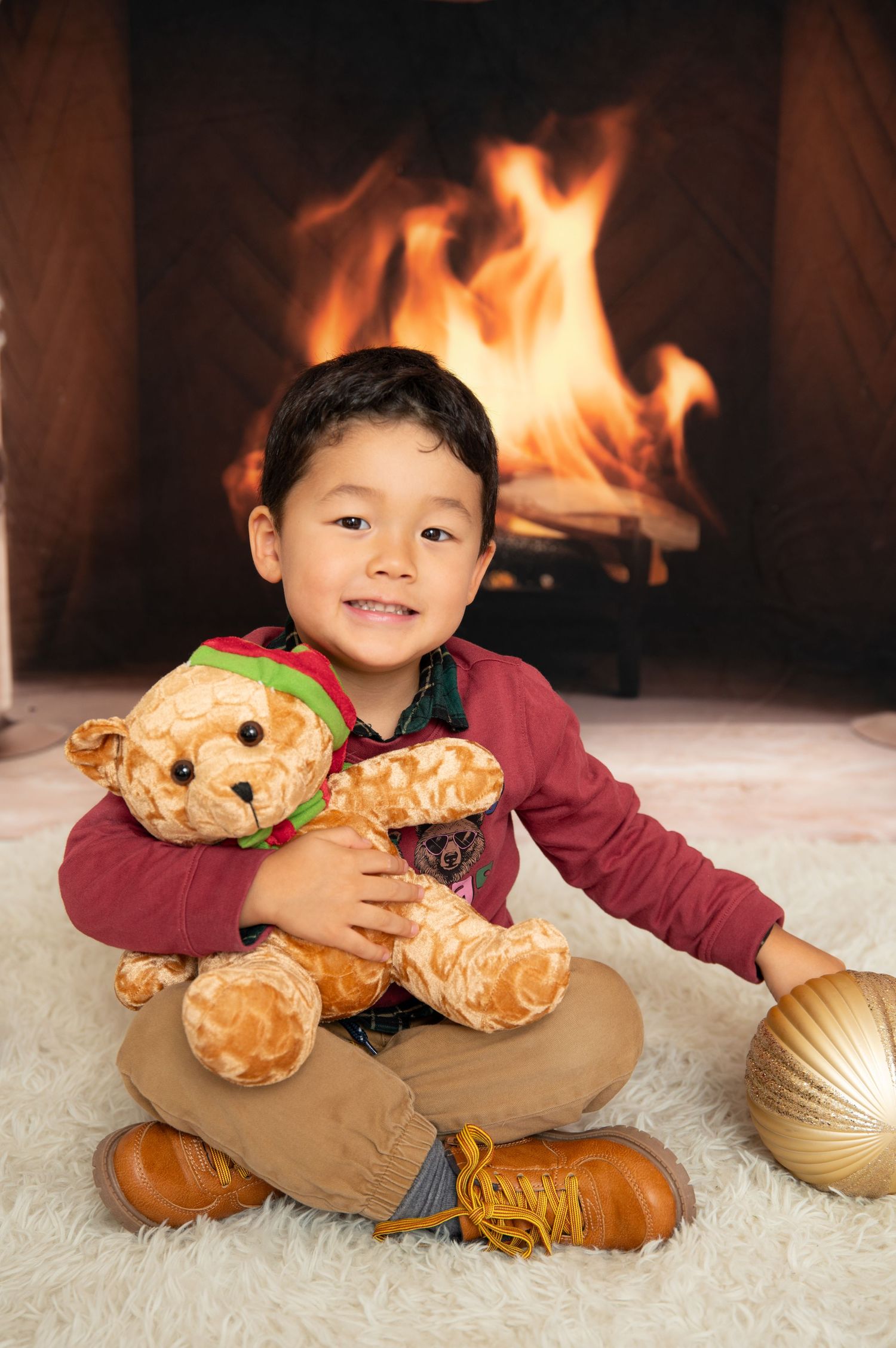 Young boy hugging a teddy bear while sitting by a cozy fireplace, wearing a red sweater and brown boots.