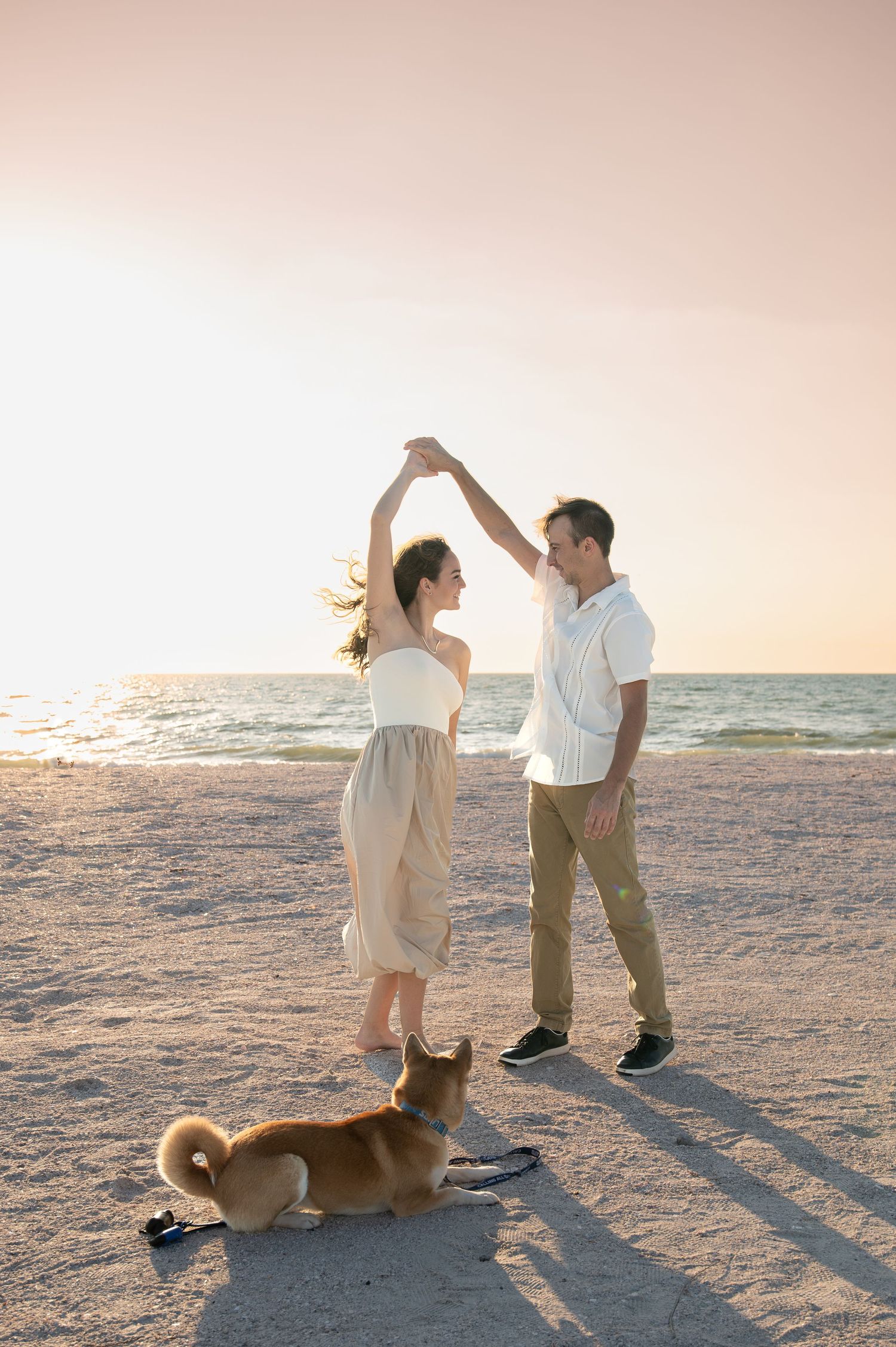 A couple dances on the beach at sunset with their dog watching nearby.
