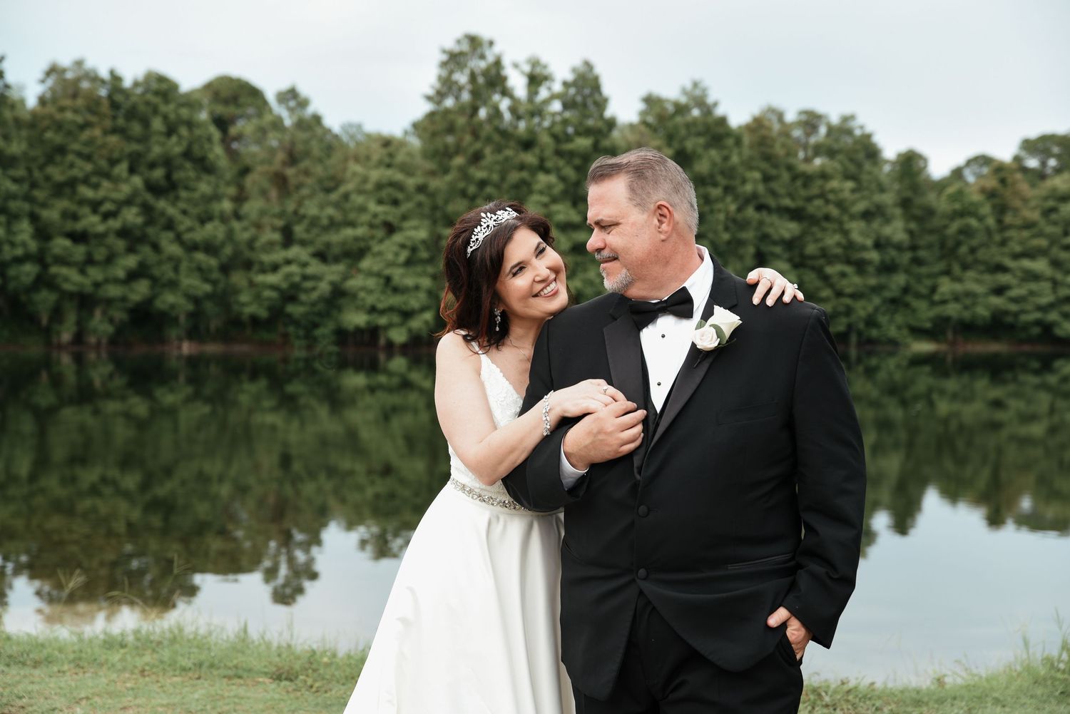 A bride in a white gown and groom in a black tuxedo embrace near a reflective lake surrounded by lush green trees.