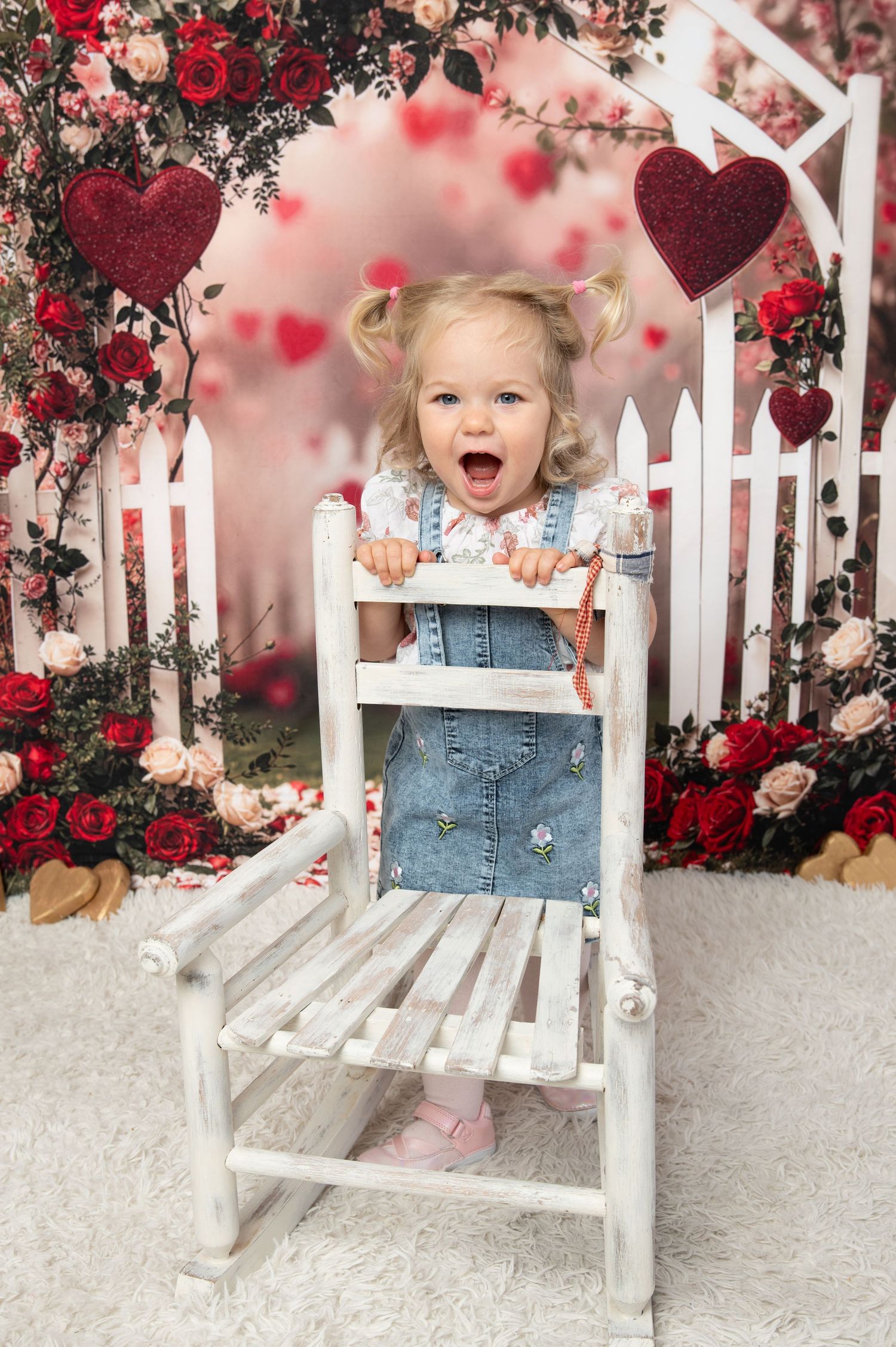 Toddler girl in denim overalls laughing behind white chair, Valentine's Day photo backdrop with roses and hearts.