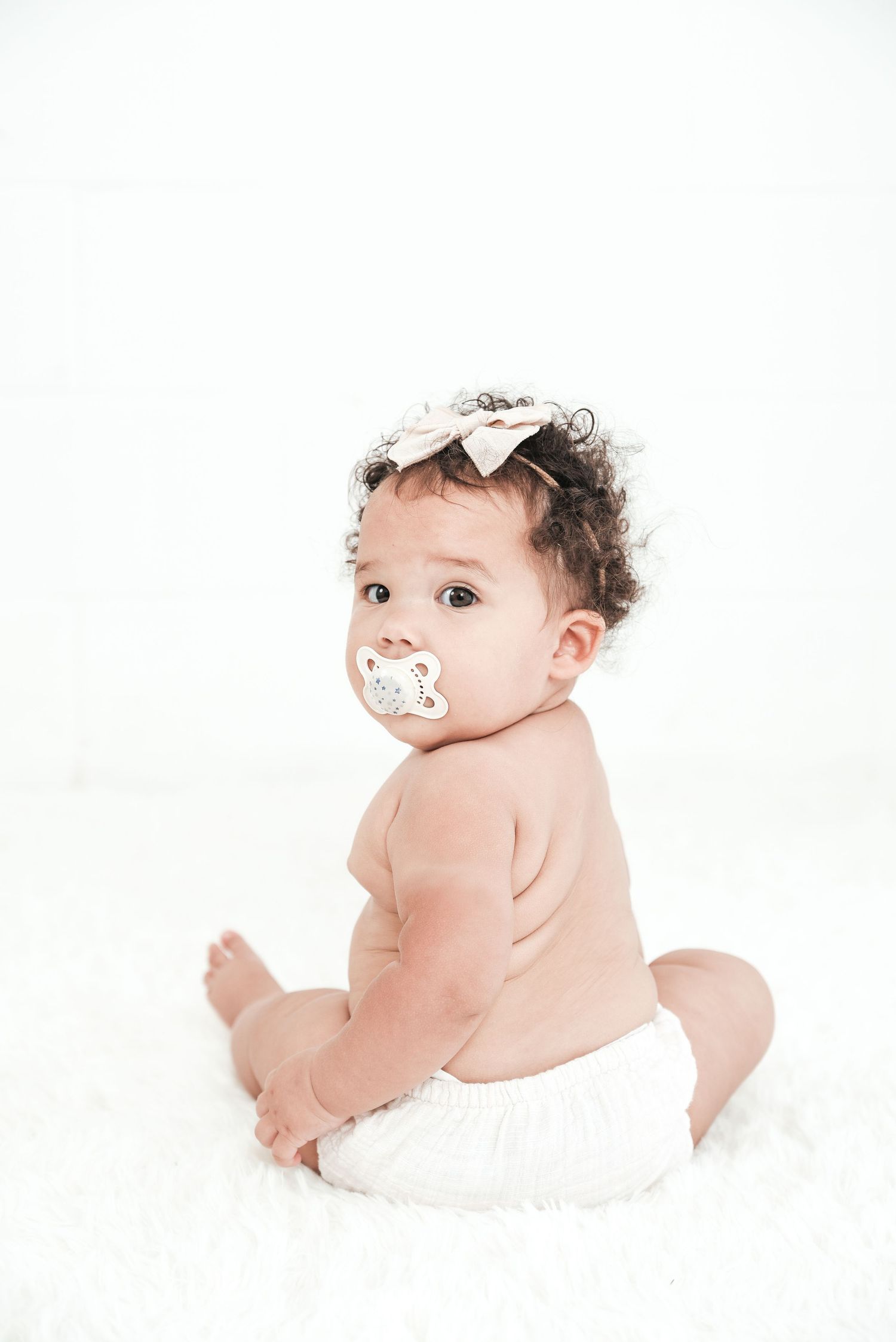 Baby wearing white bow lies on stomach on white background during portrait session.