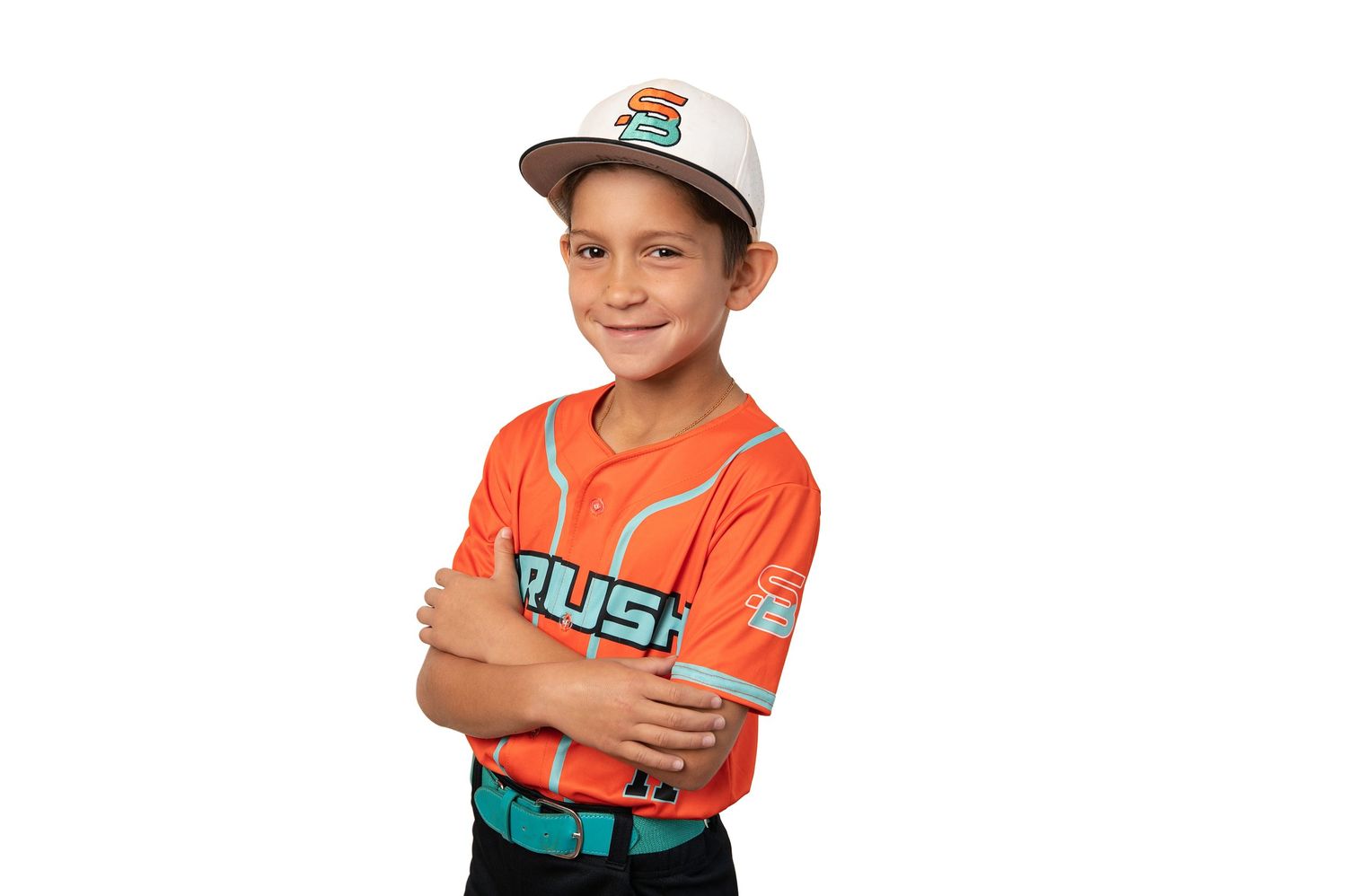Young boy in orange Rush baseball jersey and cap poses with arms crossed, smiling confidently against white background.