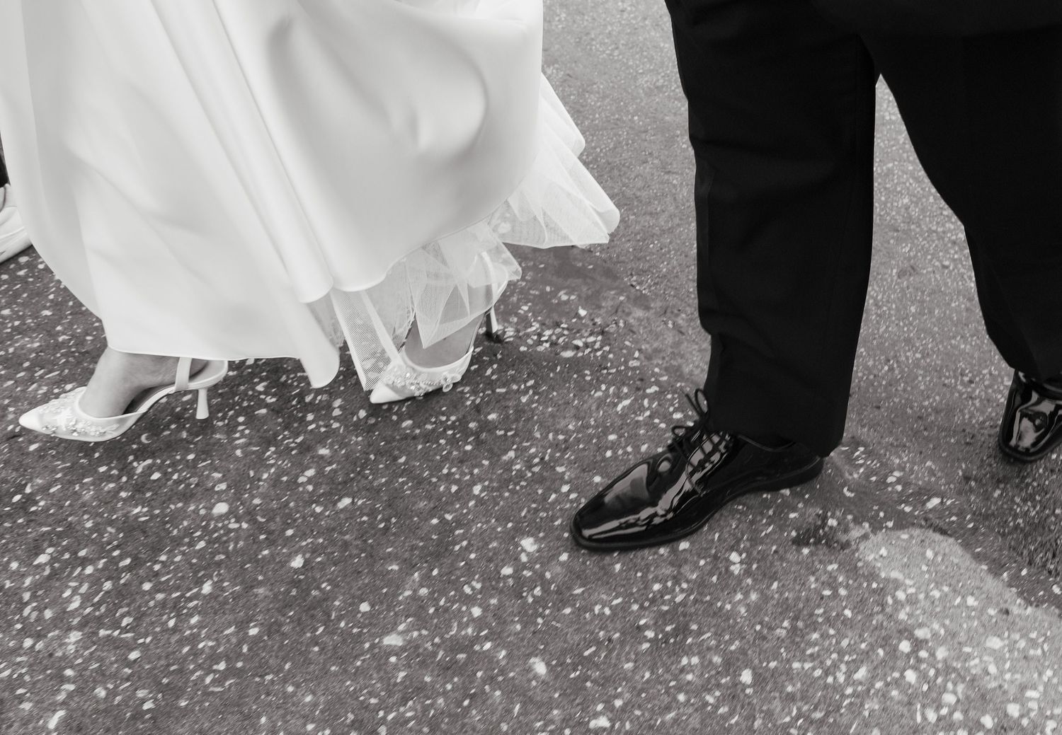 Black and white photo of bride in white gown and groom in black suit standing on pavement scattered with confetti.