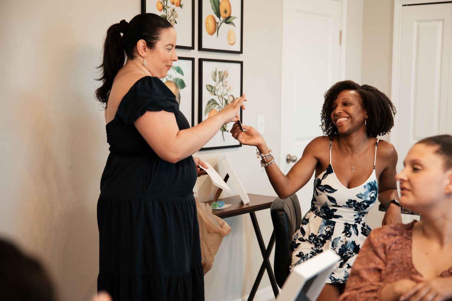 Two women laughing and exchanging gifts at an indoor social gathering, with framed botanical art on the wall.