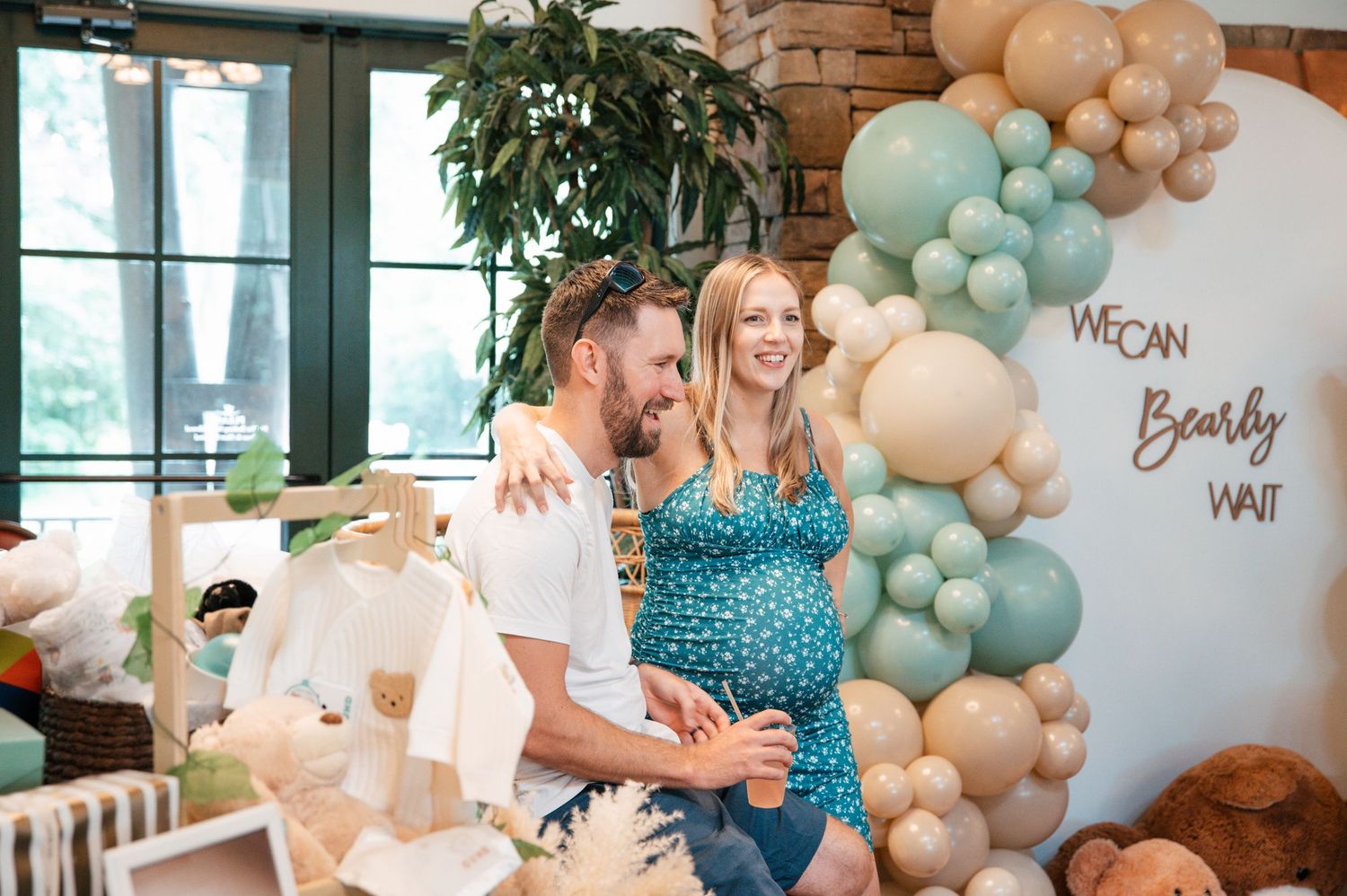 Couple at a bear-themed baby shower with balloon arch and 'We Can Bearly Wait' sign.