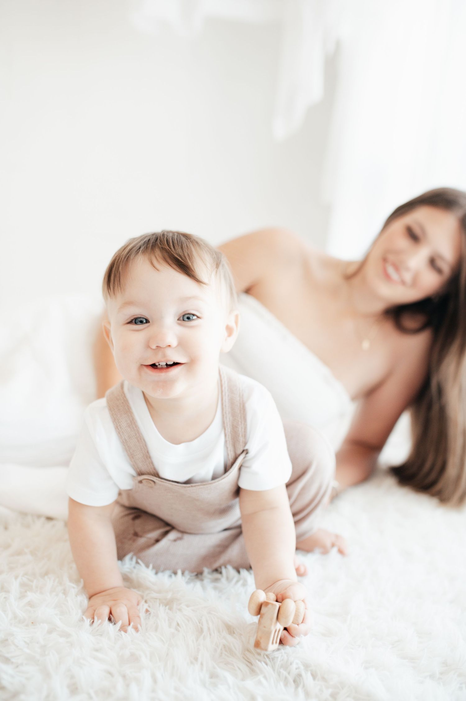 Baby in white outfit crawls on fluffy carpet while adult rests in background.