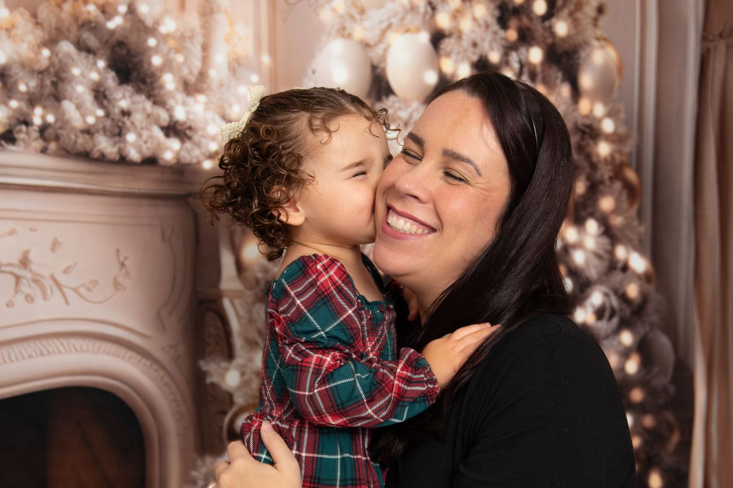 A woman and toddler share a joyful holiday moment by a decorated fireplace and Christmas tree.
