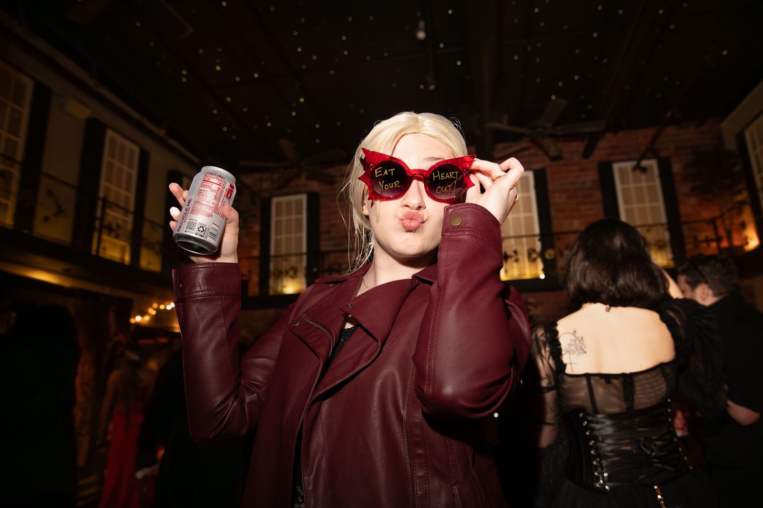 Blonde woman in red sunglasses and burgundy leather jacket holding a can at a dark, starlit indoor party.