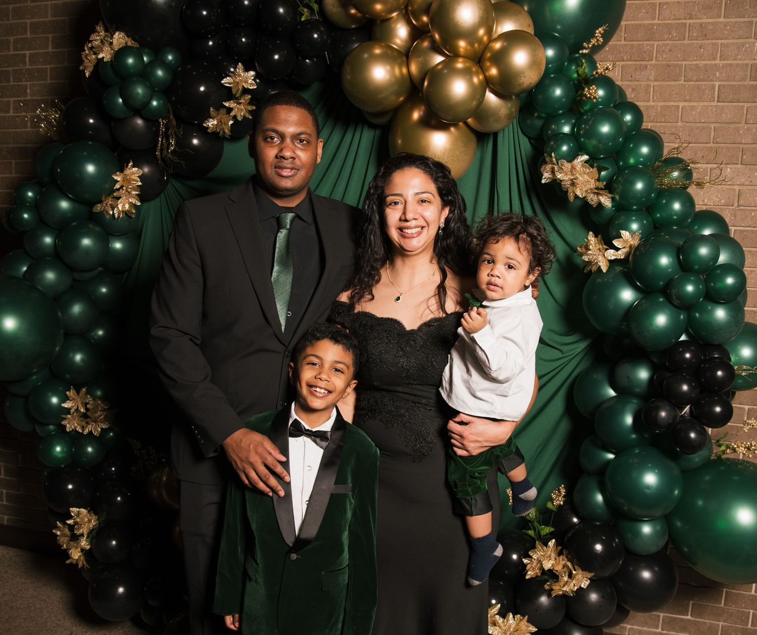 Family of four posing in front of green and gold balloon arch backdrop in festive holiday attire.