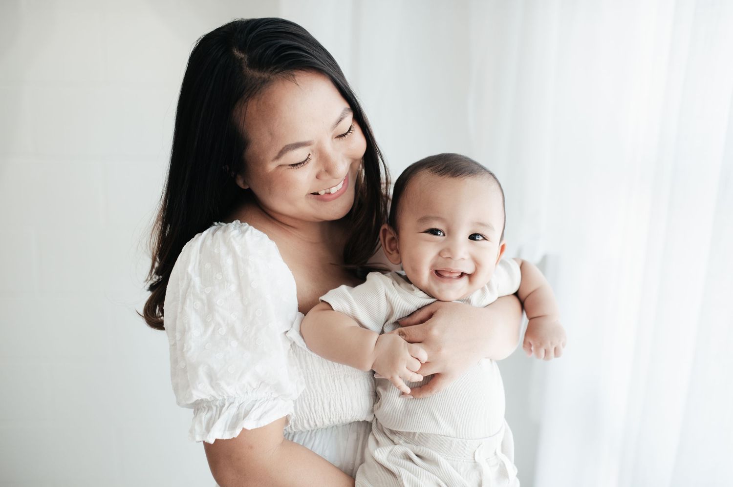 Parent and baby in white outfits sharing tender moments on white bedding.