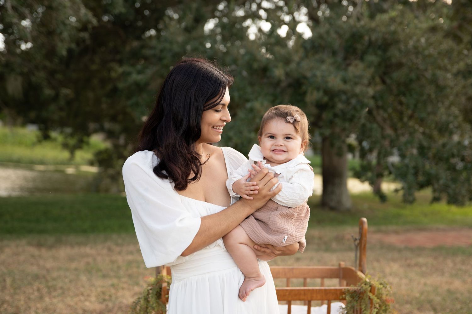 A smiling dark-haired woman in a white dress holds a happy baby girl outdoors among green trees.