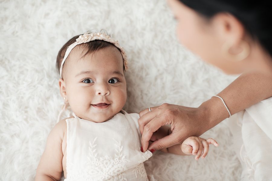 Baby in white lace dress lying on plush white carpet.