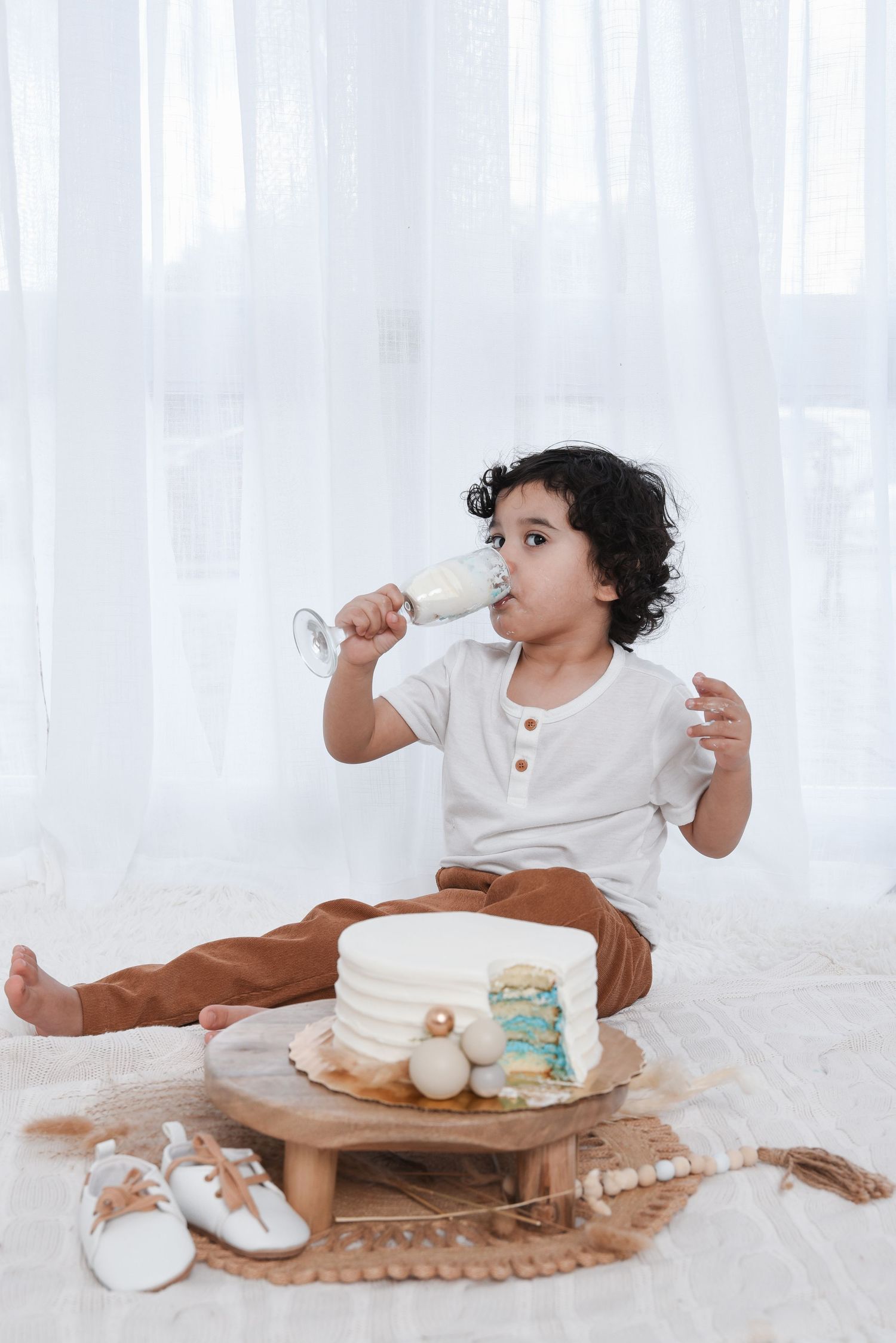 Small child in white shirt and brown pants sits on floor drinking from glass with cookies and macarons on wooden platter.