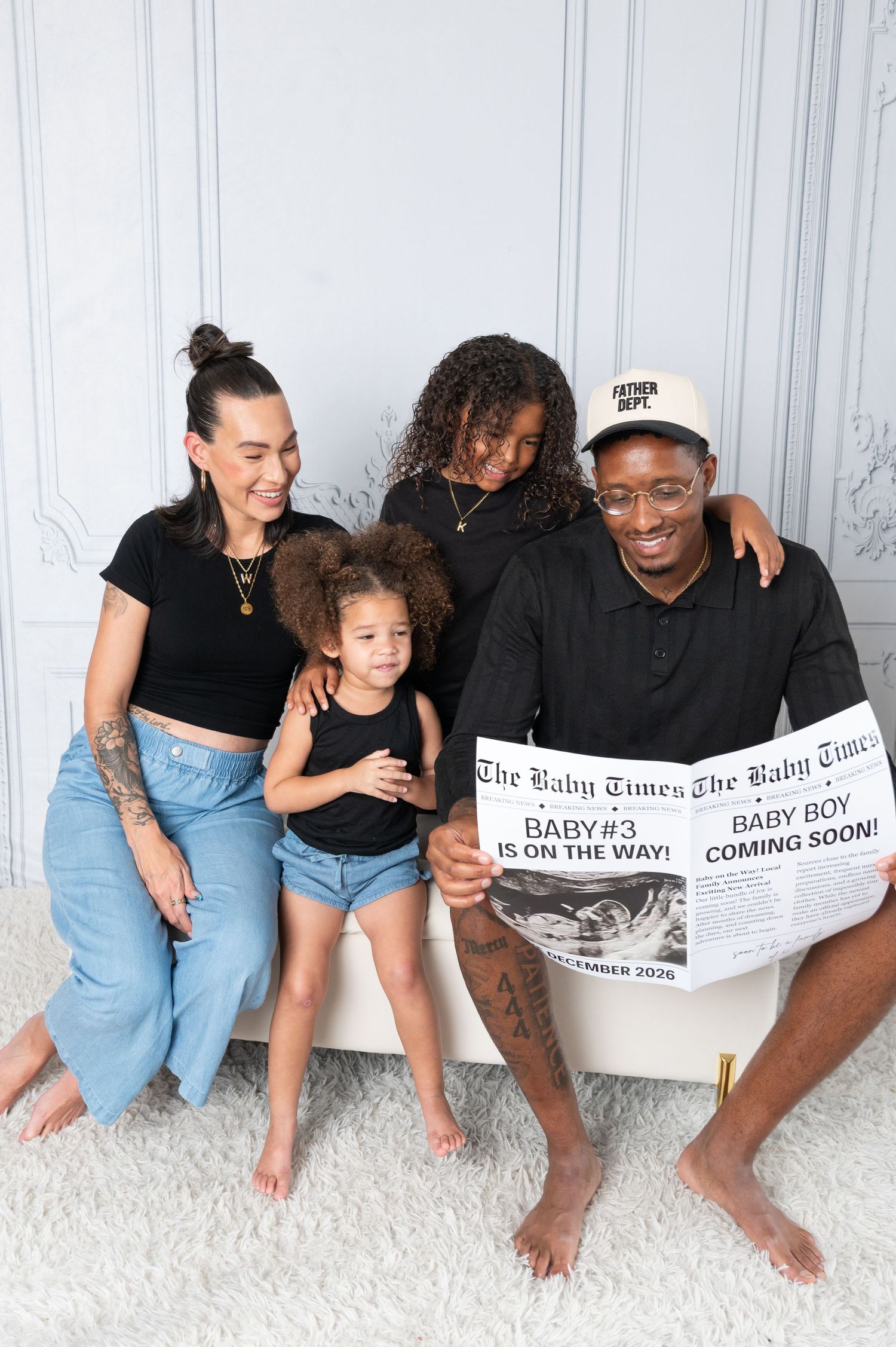 Family of four poses with a Baby Times newspaper announcing baby boy #3 coming soon in a bright studio.