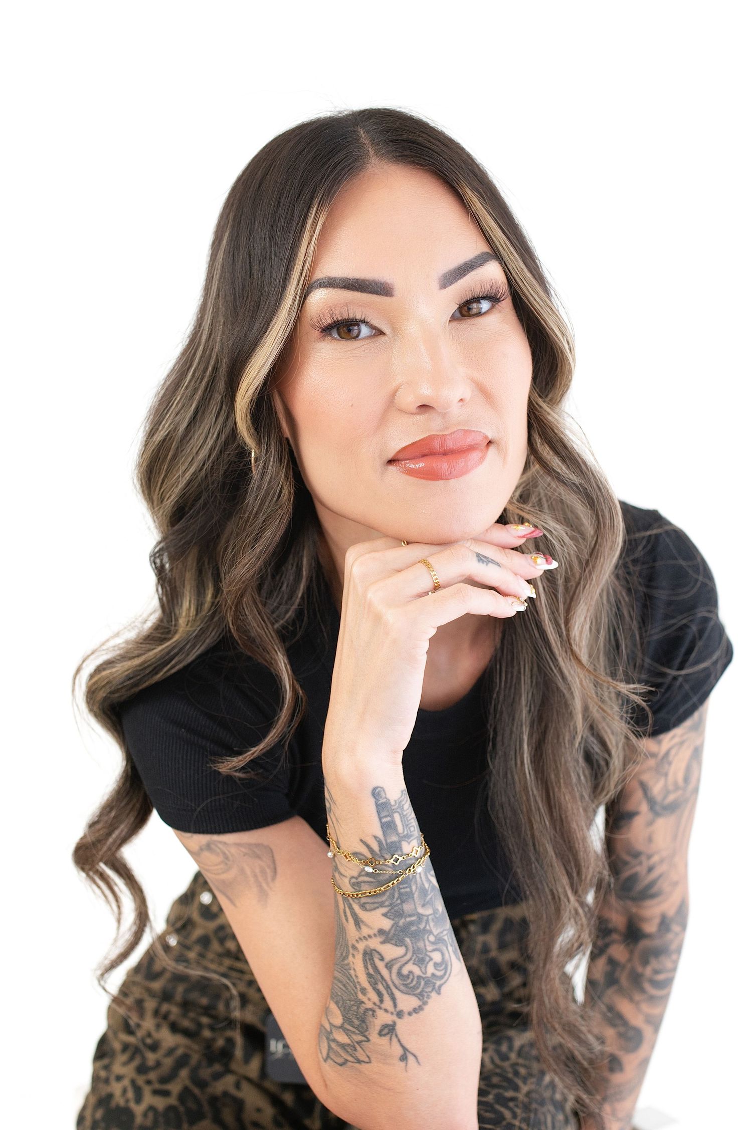 Woman with long brown hair and tattoos poses in black top against white backdrop.