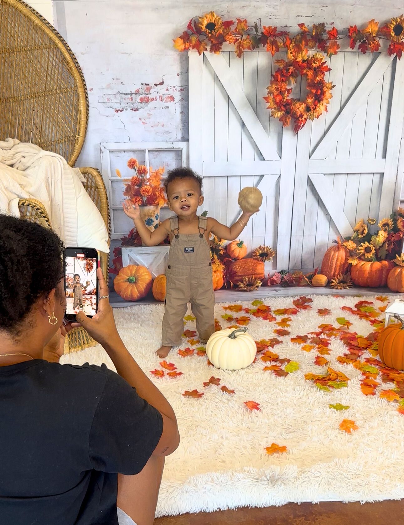 A fall-themed photo session setup with white barn doors, orange wreaths, and pumpkins while someone takes a photo.