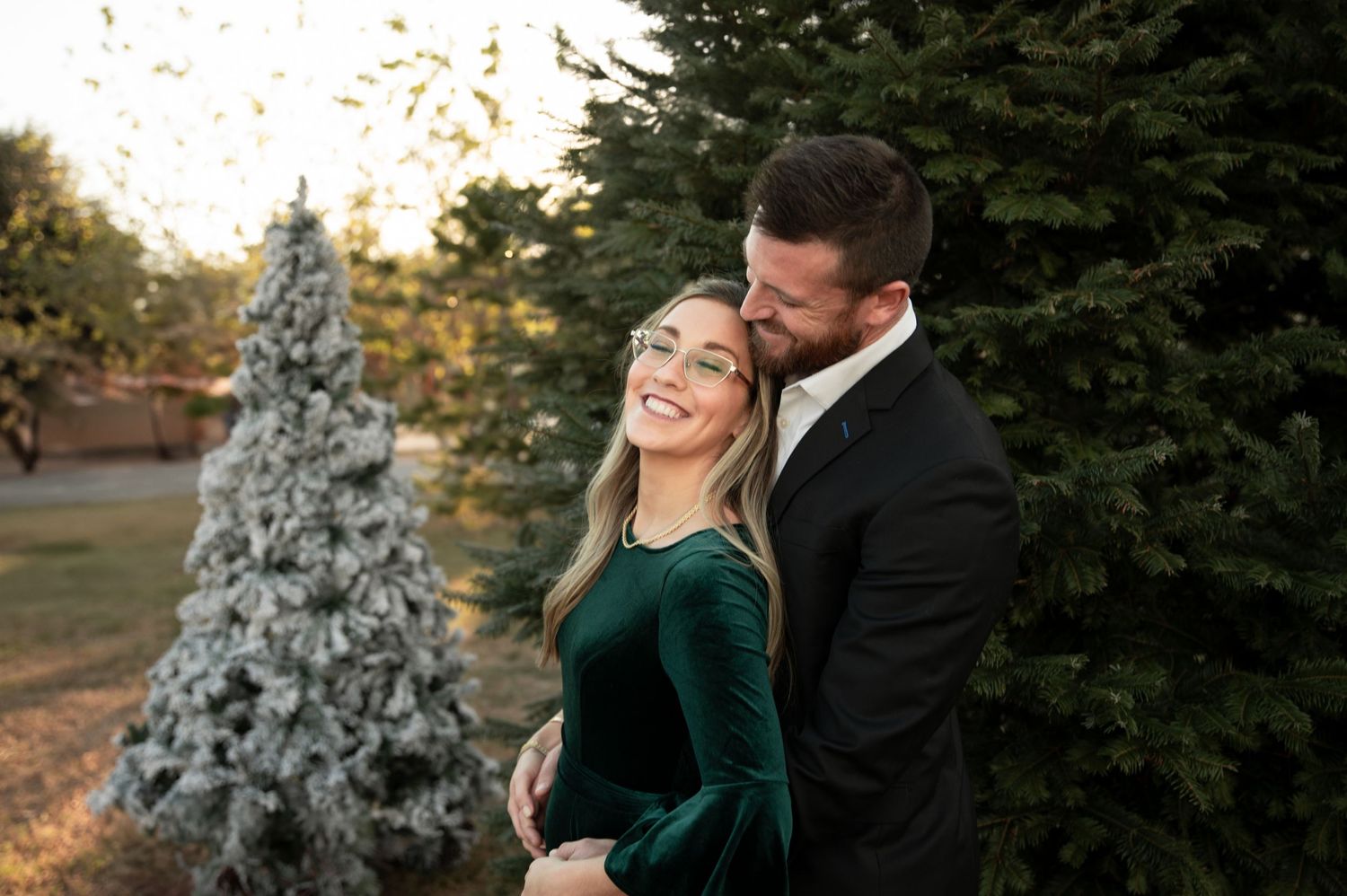 Happy couple embracing outdoors near snow-dusted Christmas trees, woman in green velvet dress, man in dark suit.
