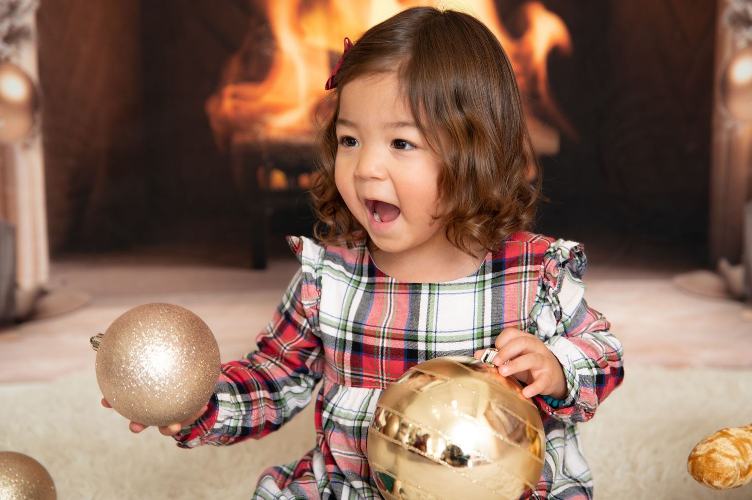 Young girl in plaid dress holding gold Christmas ornaments by a cozy fireplace.