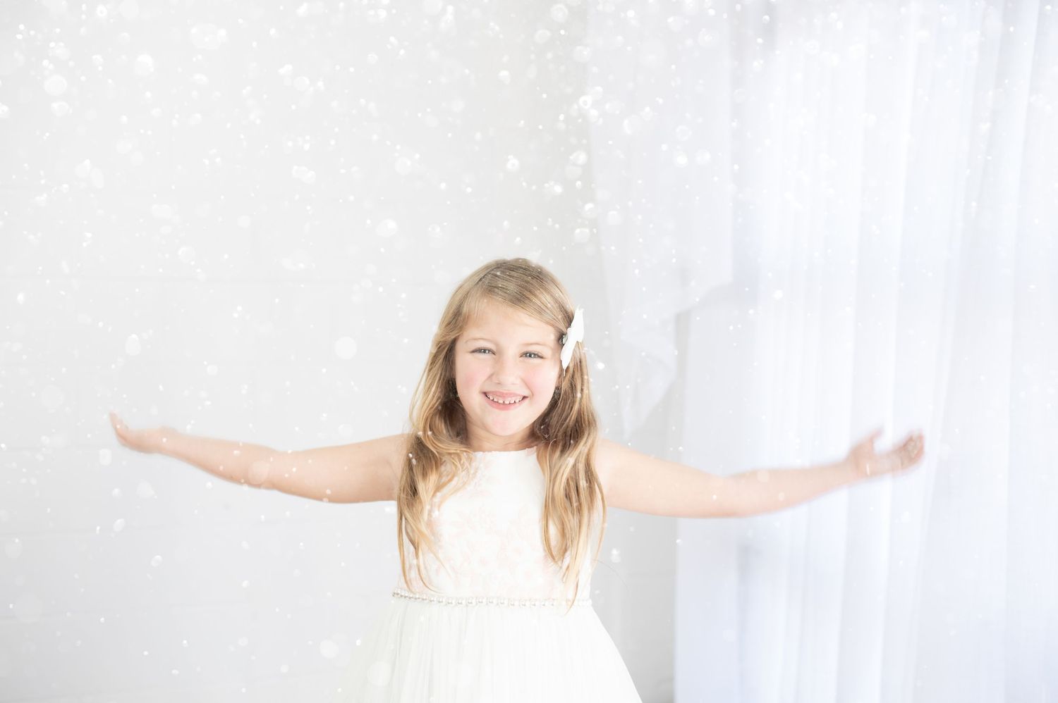 A joyful young girl in a white dress spreads her arms wide, surrounded by sparkling snowflakes in a bright studio.