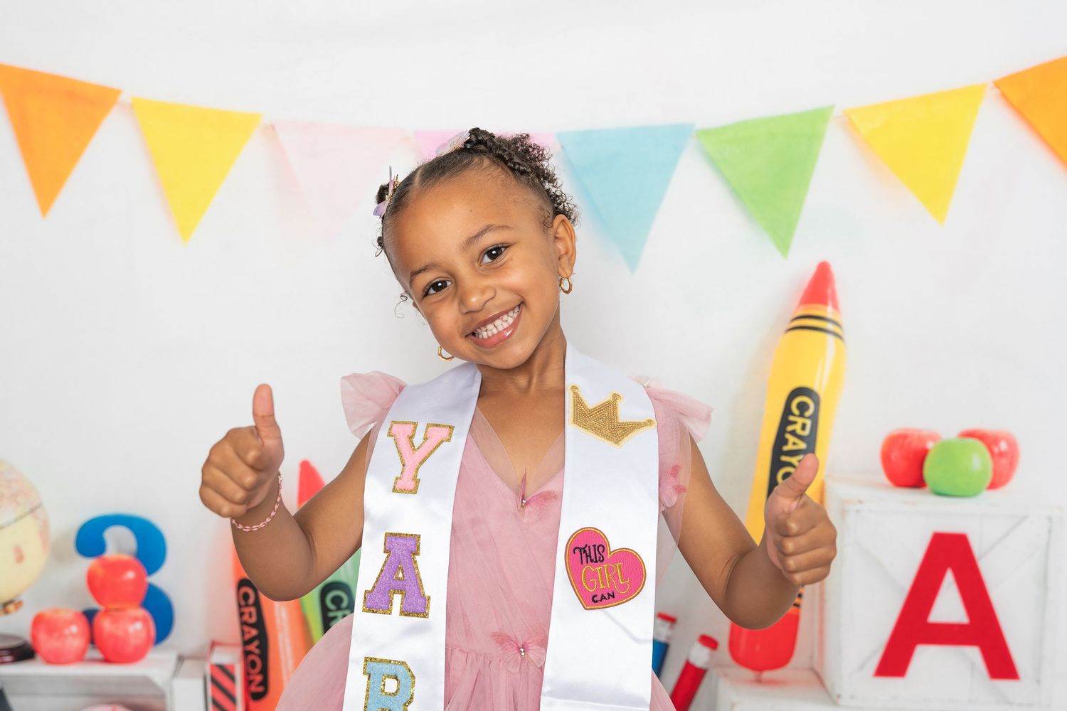 Young girl in a sash holding a giant crayon prop, giving thumbs up at a colorful back-to-school photo shoot.