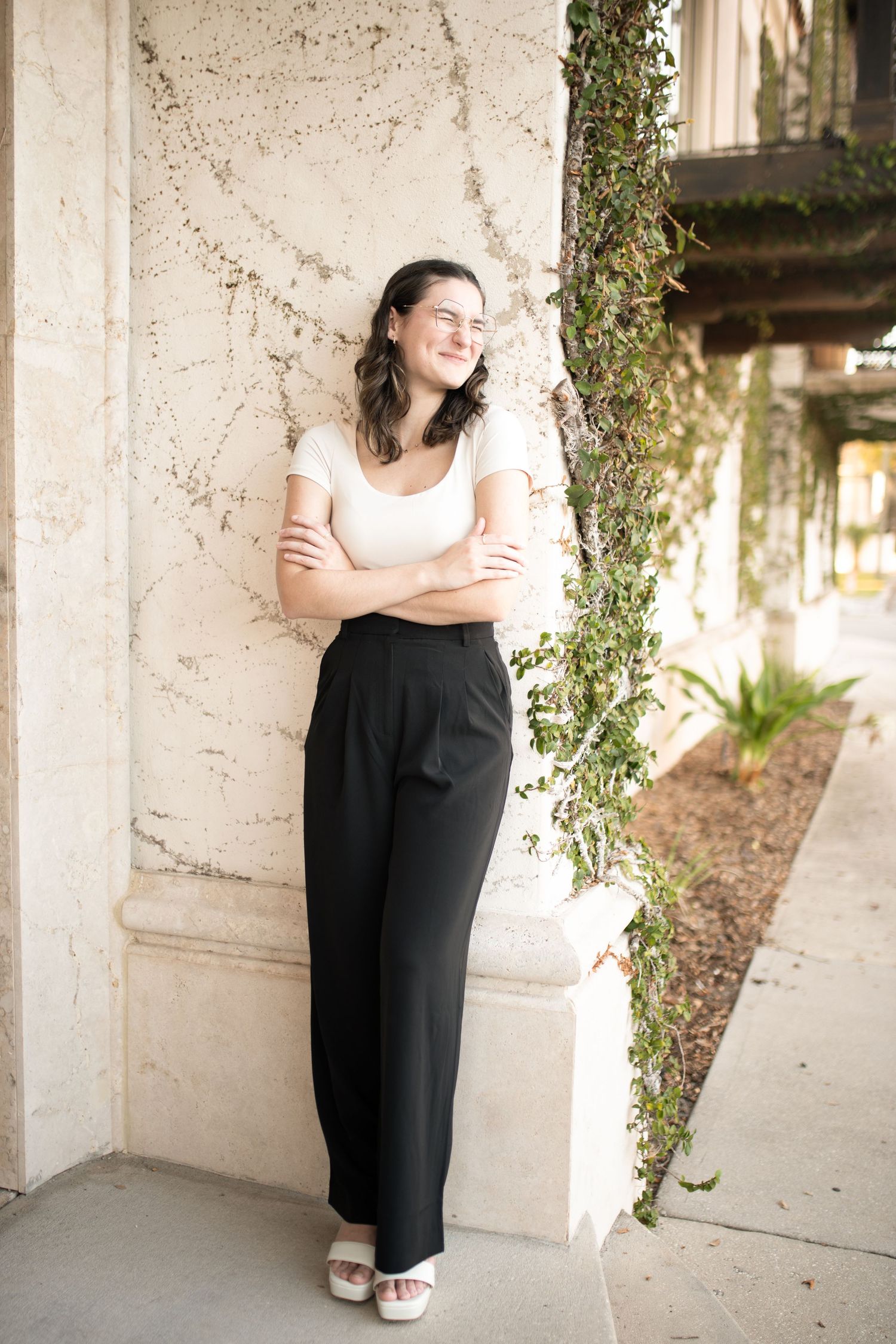 Woman in white top and black pants standing by outdoor stone column with vines.