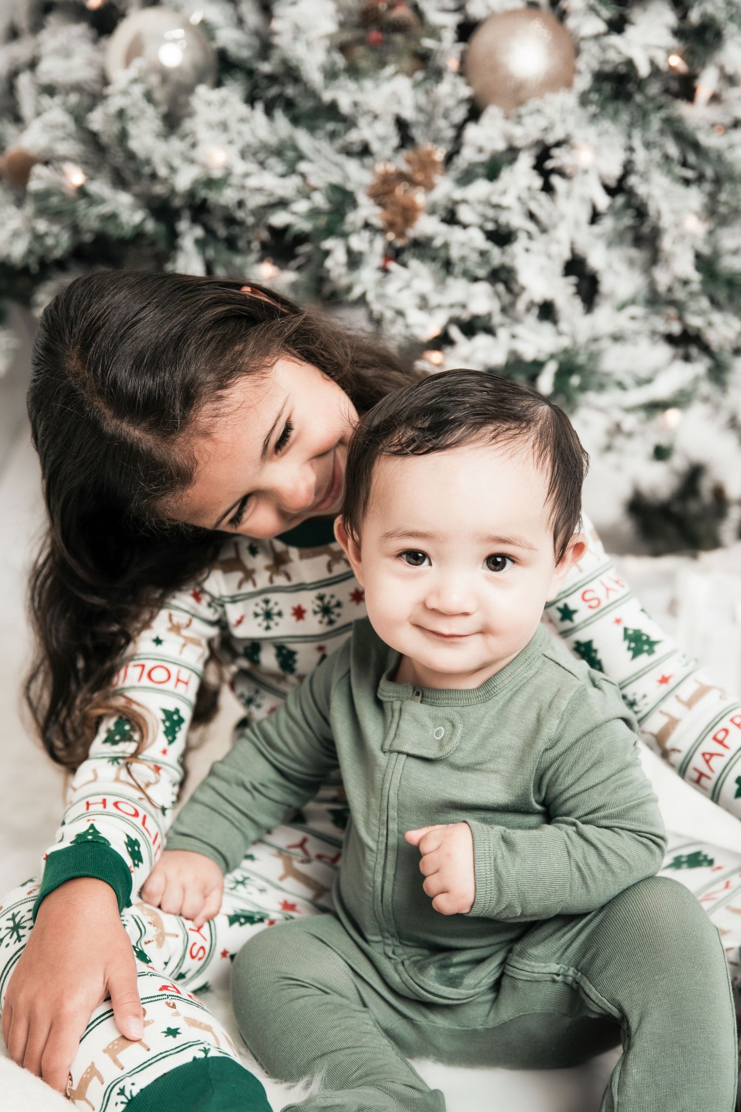 Sweet holiday moment captured in front of a snowy Christmas tree with festive decorations and matching outfits.