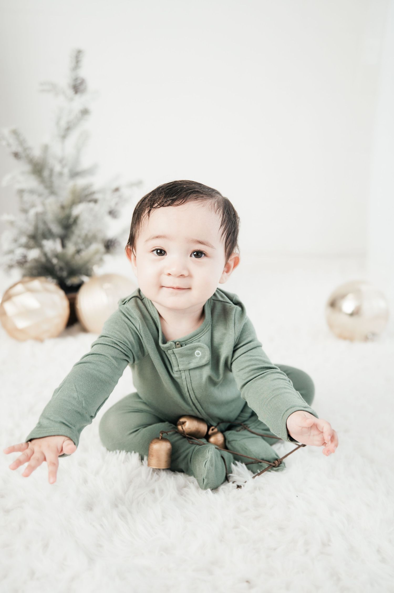 Baby in sage green outfit crawls and plays on white fluffy rug with winter holiday decor in background.