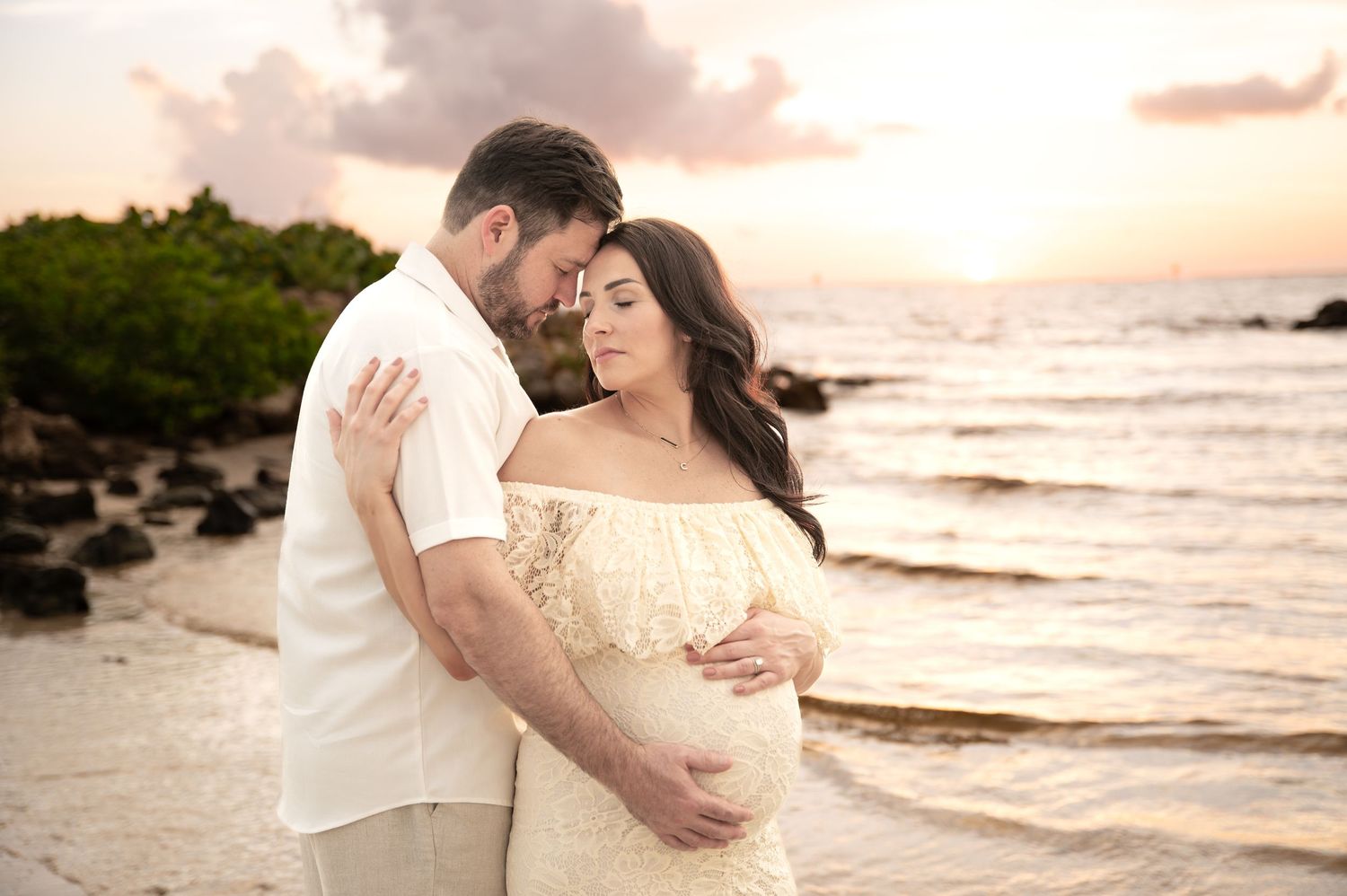 Couple sharing tender moment on beach during sunset with ocean waves behind them.