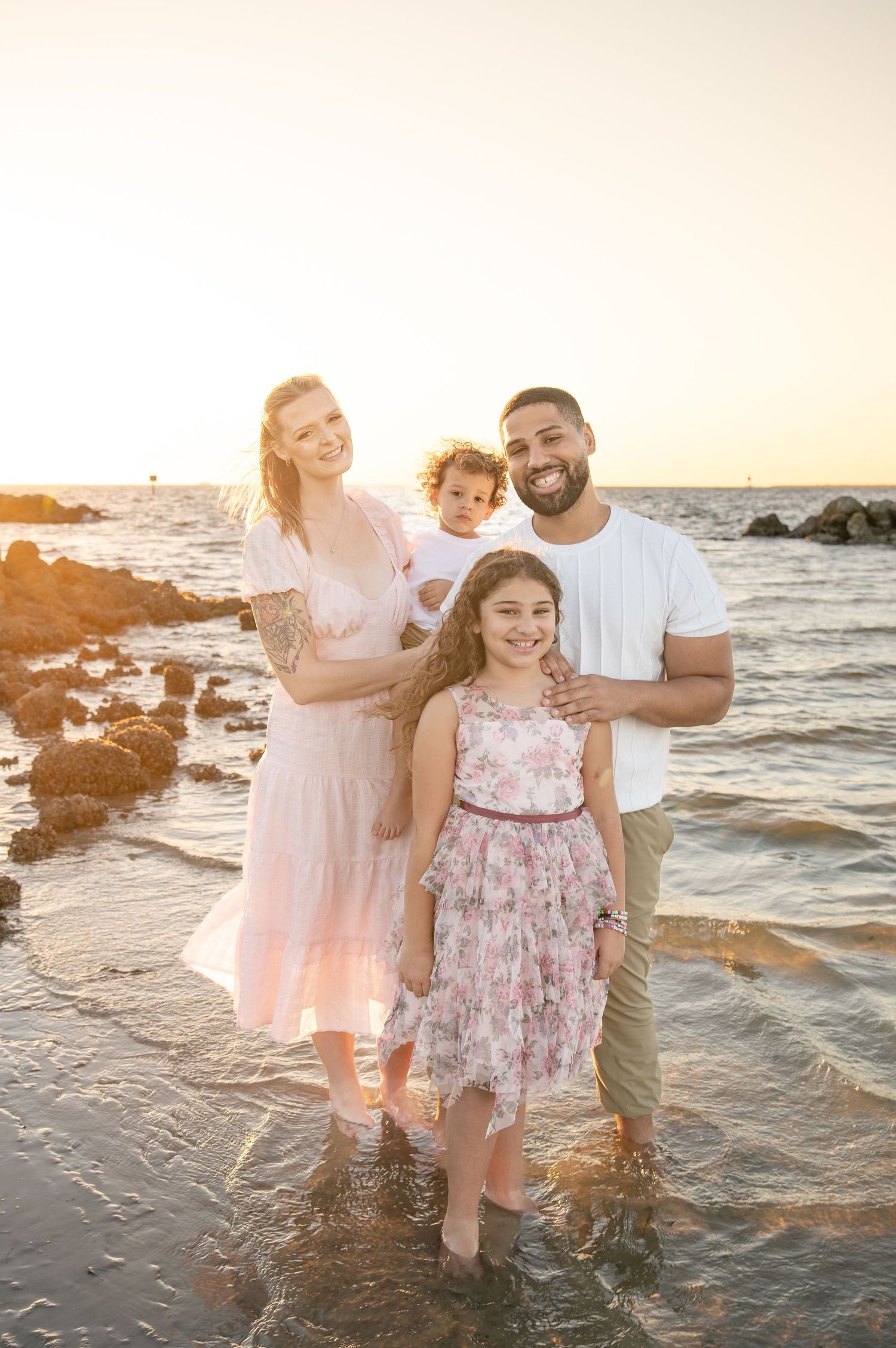 Family of four posing on a rocky beach at golden hour, wearing pastel outfits, smiling at the camera.