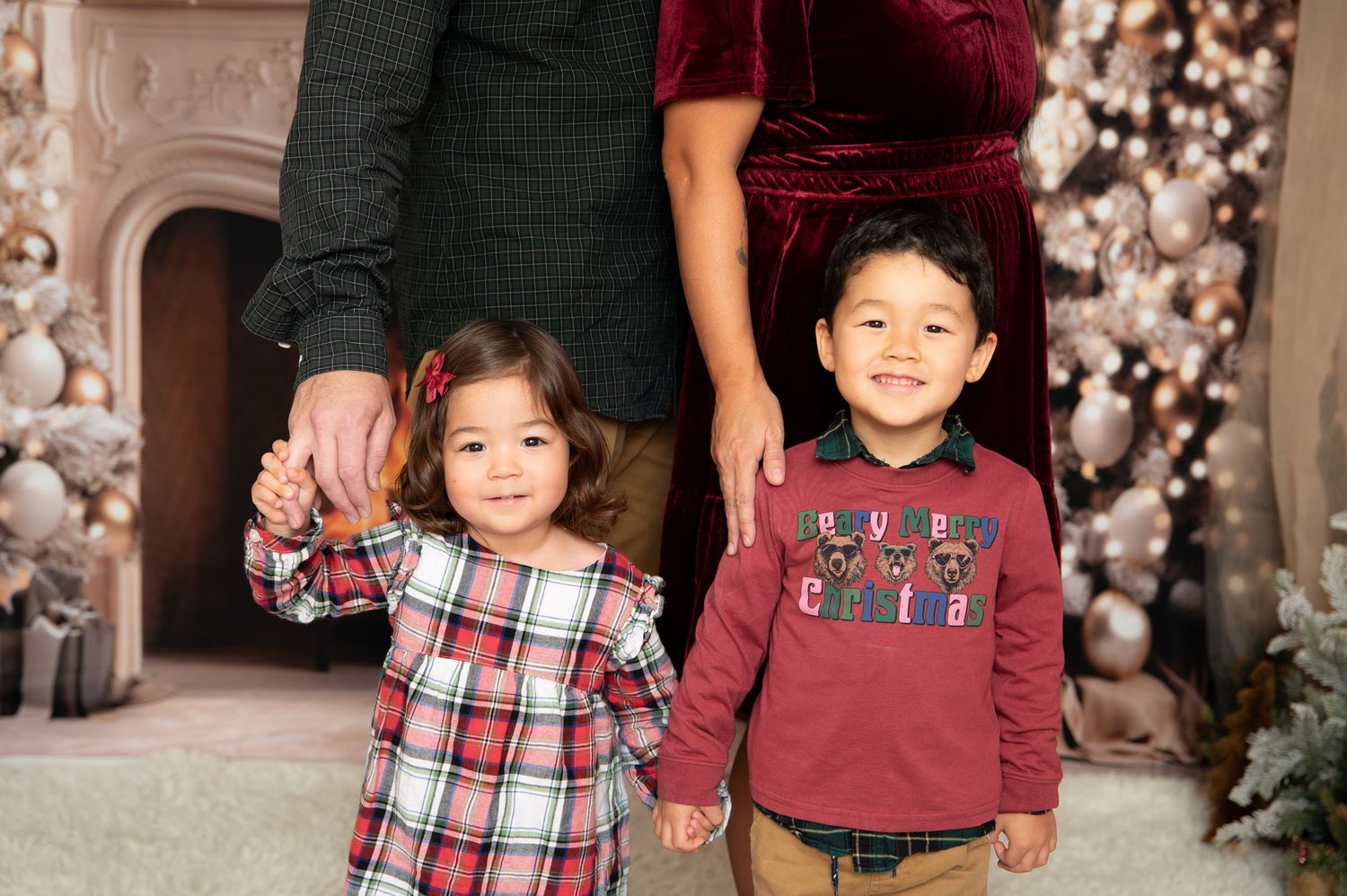 Two young children in holiday outfits smile together in front of a decorated Christmas tree and fireplace backdrop.