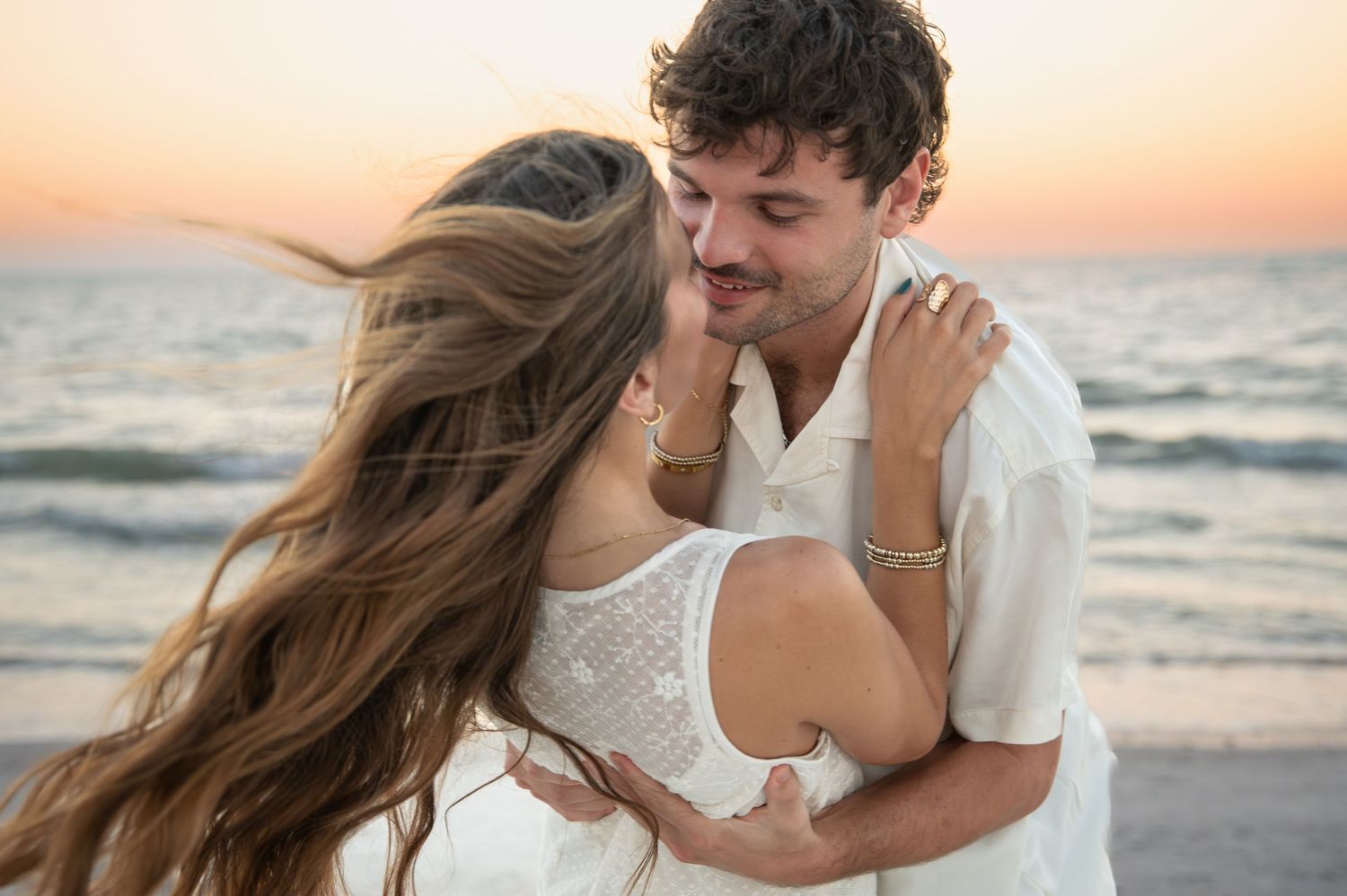 Couple in white outfits embracing on beach at sunset.