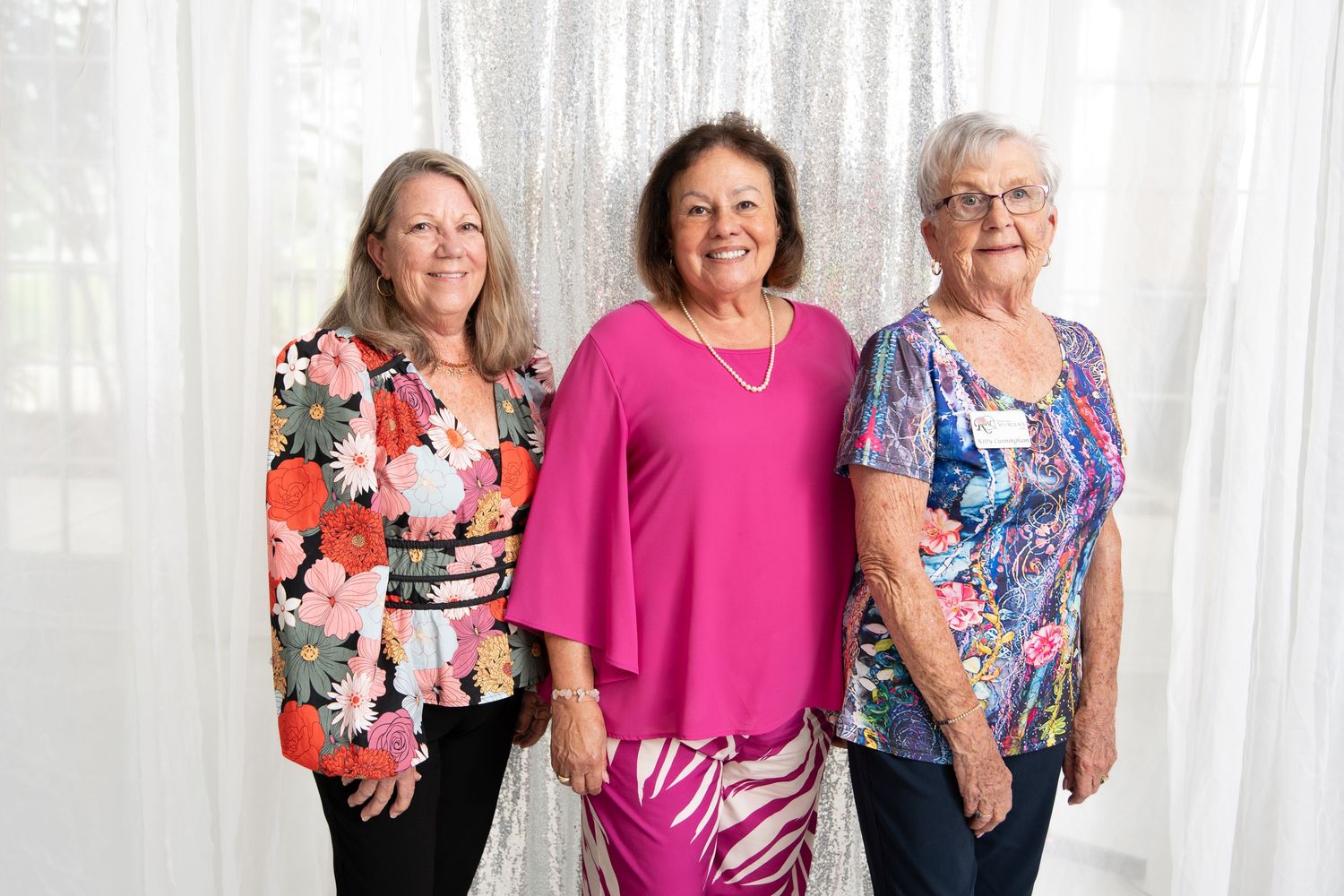 Three smiling women posing together in colorful outfits against a silver sequin backdrop.