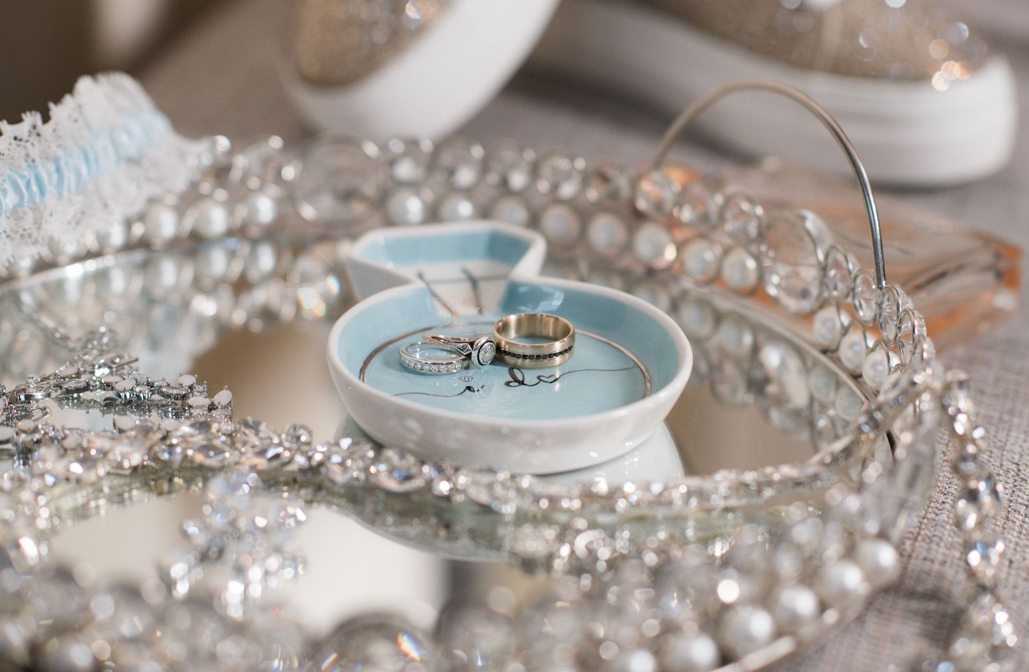 Wedding rings displayed on a light blue dish surrounded by sparkling crystal beads and silver jewelry on a mirrored tray.