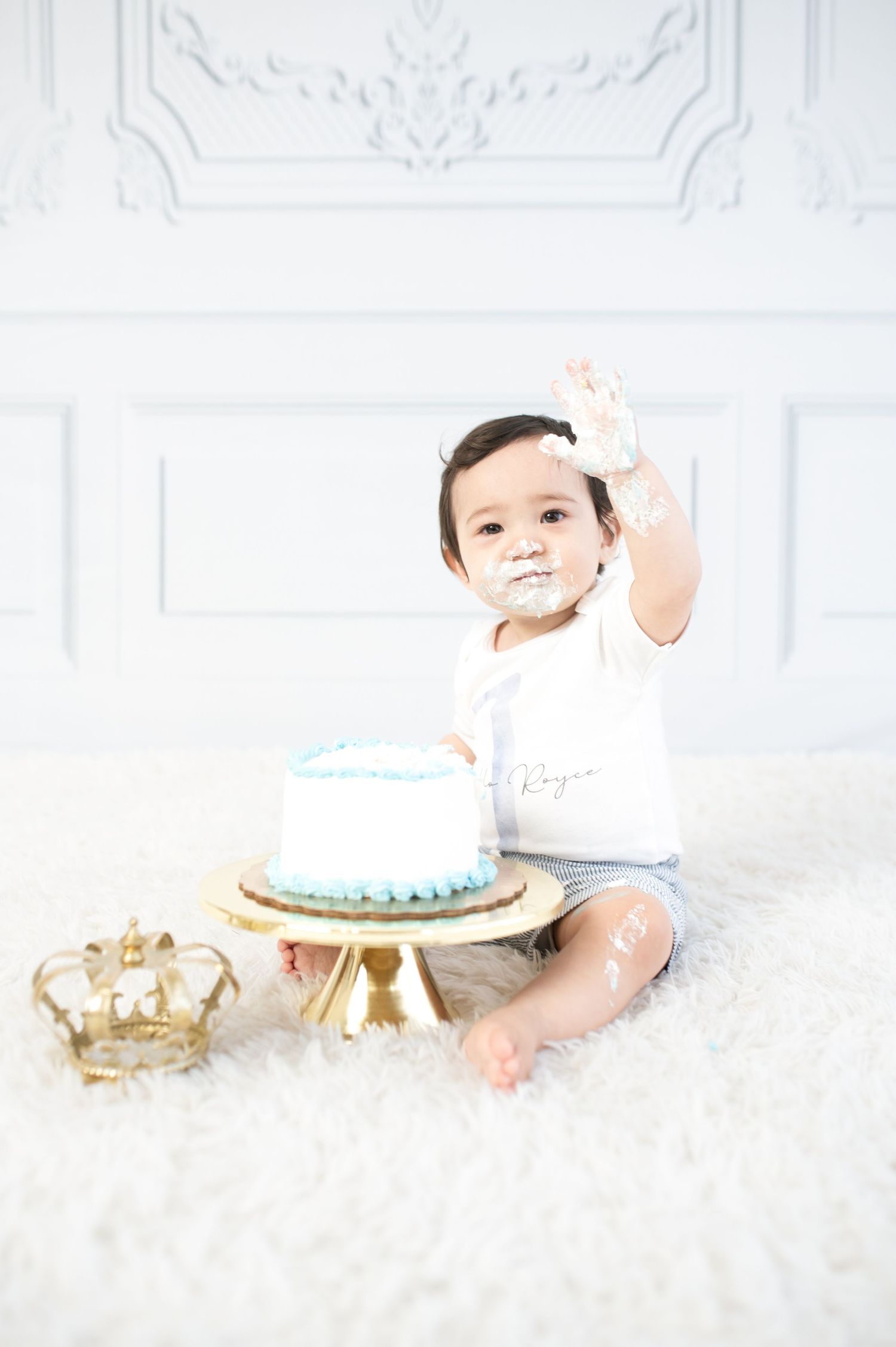 Baby in white outfit reaches toward decorated cake during first birthday photoshoot on white carpet.