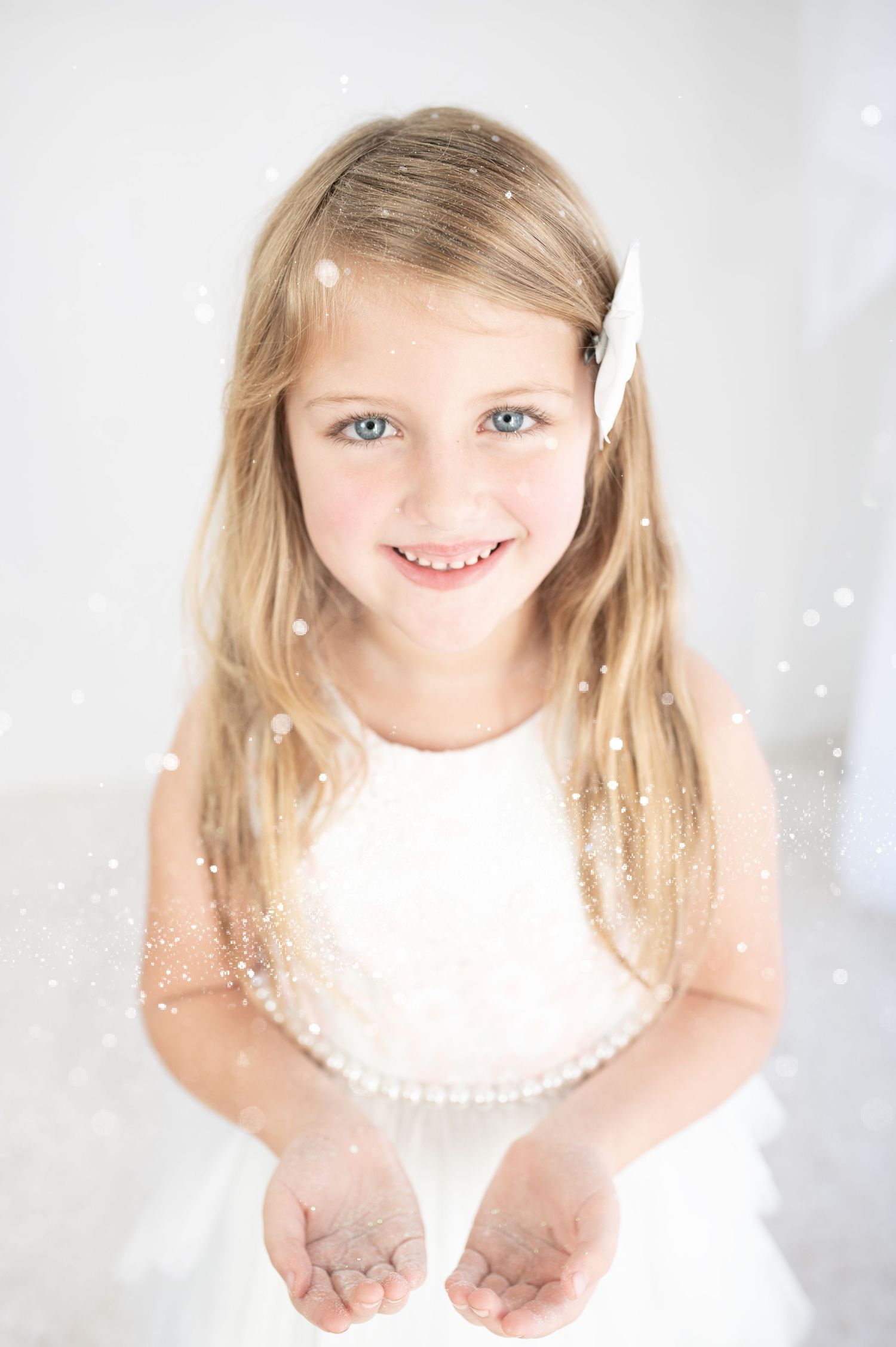 Young blonde girl in white dress holding out hands catching snowflakes, smiling with blue eyes and hair bow.